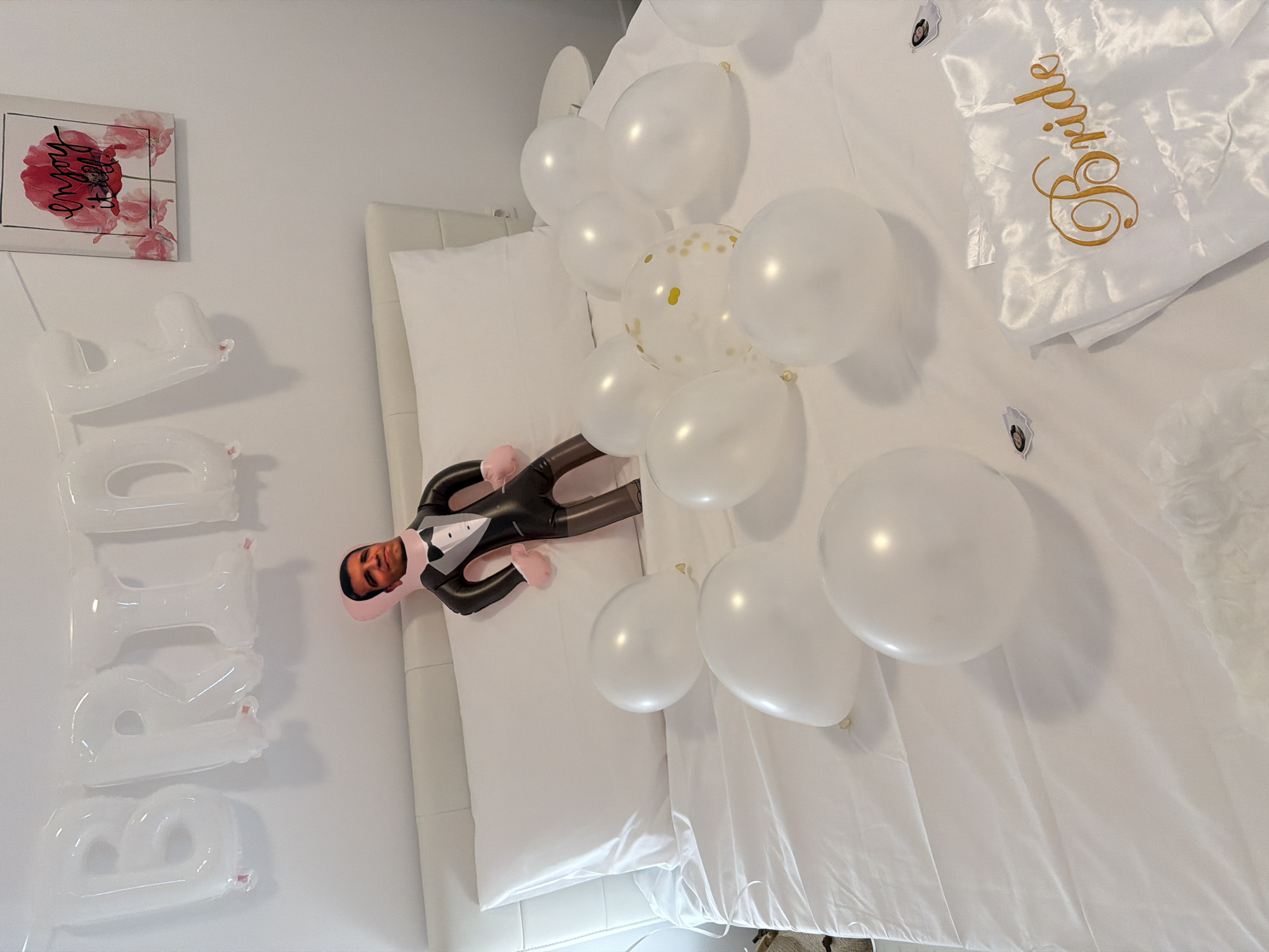 A bridal bedroom is decorated for a bachelorette party. A white inflatable "BRIDE" balloon banner is hung on the wall above a bed with white linens. A small framed picture of red poppies with the text "enjoy it all" is also on the wall. A blow-up doll dressed in a tuxedo is lying on the bed amidst a scattering of white and gold confetti-filled balloons. A white satin robe with "Bride" embroidered in gold script is folded on the bed in the foreground. There are no people in the image. The location is not determinable from the image.