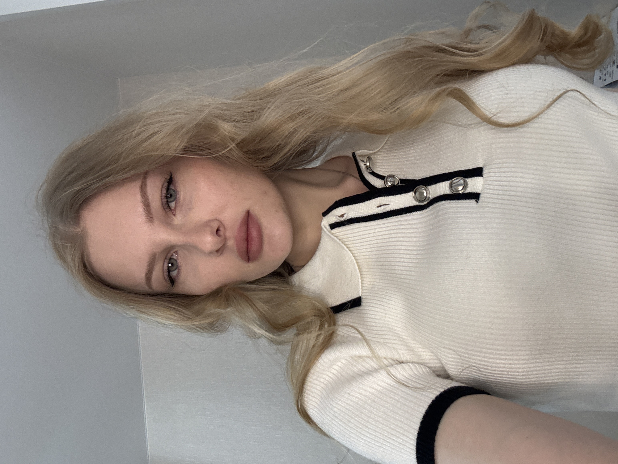 A woman with long blonde hair is wearing a white, ribbed top with a black stripe and silver buttons. The background is a plain, light grey wall. The photo was taken indoors. The location in the city of Perm, Russia is not discernable from the image.