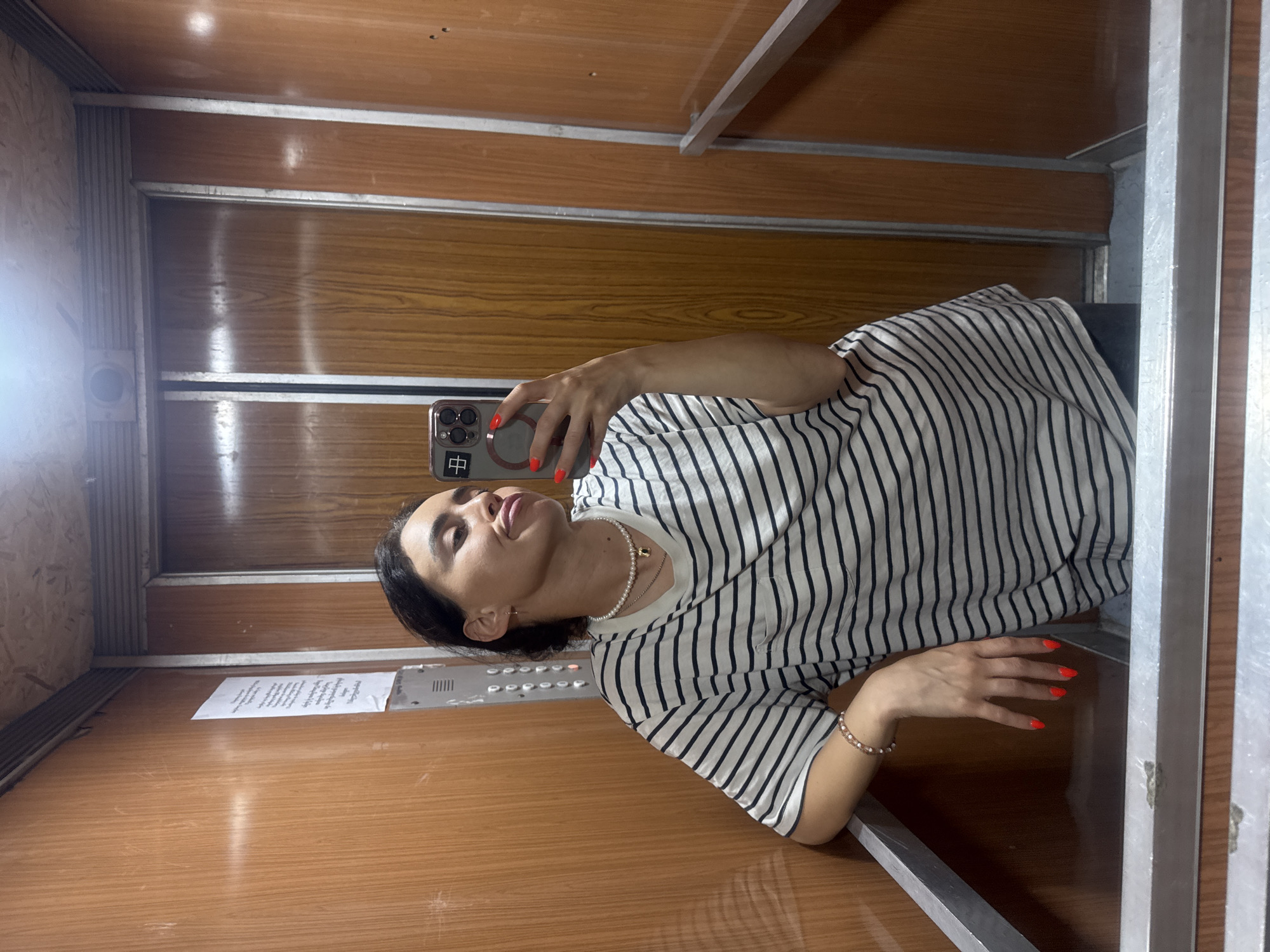 A young woman with dark hair, wearing a black and white striped t-shirt, a pearl necklace, and a bracelet, takes a selfie in an elevator. Her fingernails are painted bright red, and she is puckering her lips. The elevator has wood-paneled walls and a metal handrail. A notice is affixed to the wall. The image appears to be taken in Tbilisi, Georgia, based on the context provided.