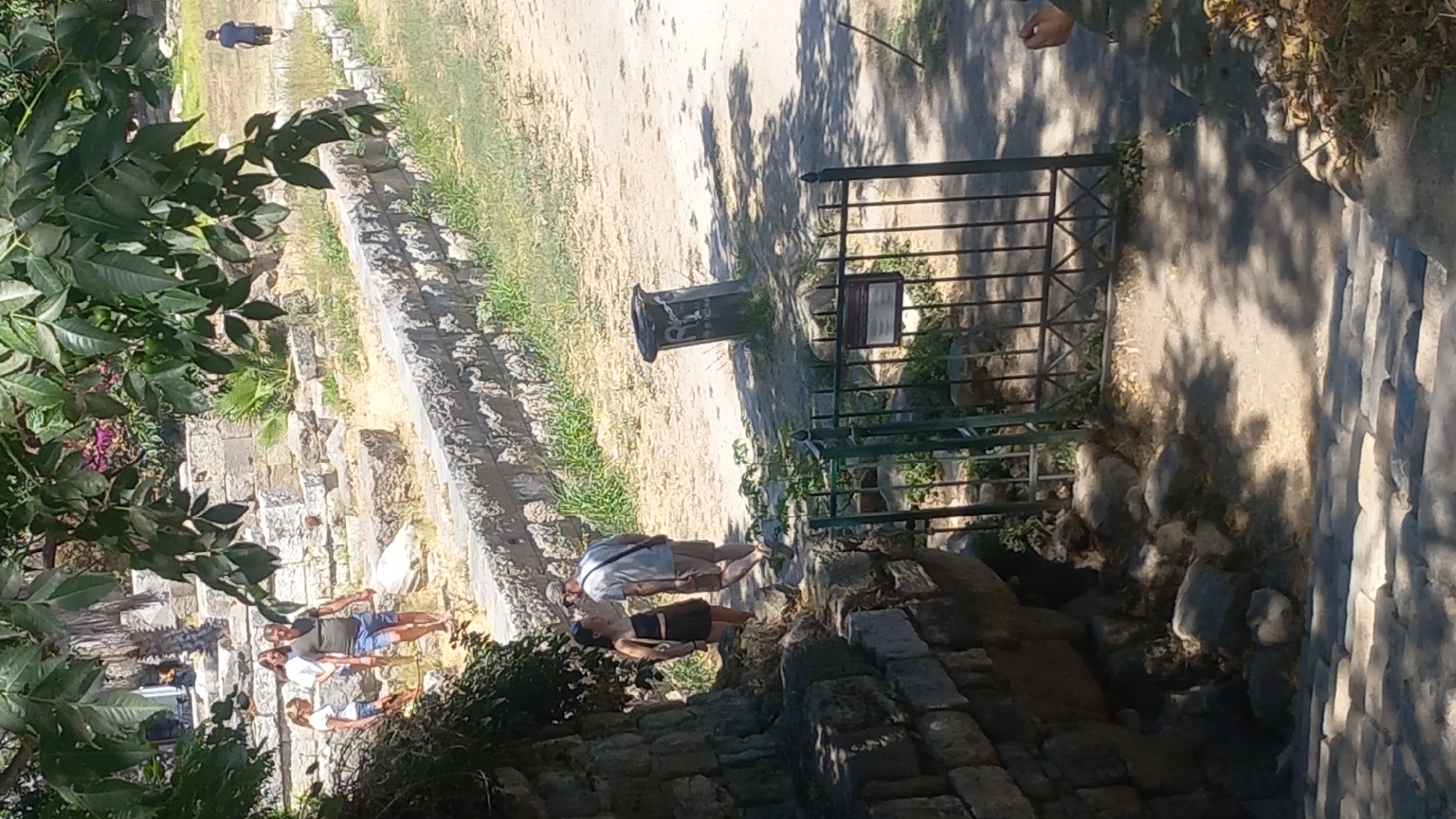 In Kos, Greece, on a sunny day, two individuals, a man and a woman, are walking side-by-side on a dirt path near ancient stone ruins. The man, wearing a light-colored t-shirt and shorts, is looking towards the woman, who is in a dark top and skirt. Behind them, a metal gate stands open, with a sign attached to it. Further in the background, more stone structures and scattered vegetation are visible, along with another person walking away from the camera. The foreground shows a stone pathway leading down, with dappled sunlight and shadows cast by overhead foliage.