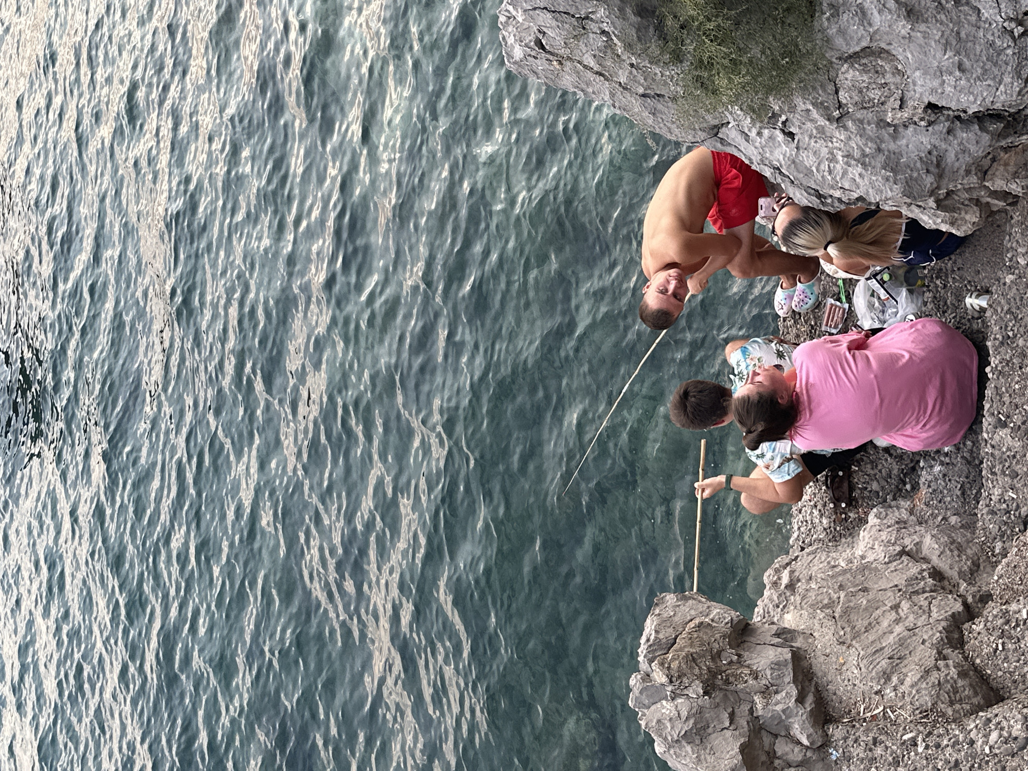 A group of four people are fishing by the water's edge on a rocky shore in Budva, Montenegro. Two individuals are seated on the rocks, facing the water. One, wearing a pink shirt, is looking away from the camera. The other, in a patterned shirt, is holding a fishing rod. A man in red swim trunks is leaning against a rock, looking at the camera. Behind him, a woman with blonde hair tied in a ponytail is looking down at items on the ground. The water is clear and blue-green, with small waves. The shore is covered in pebbles and larger rocks. There are some items scattered on the ground, including what appears to be food and a small silver container.