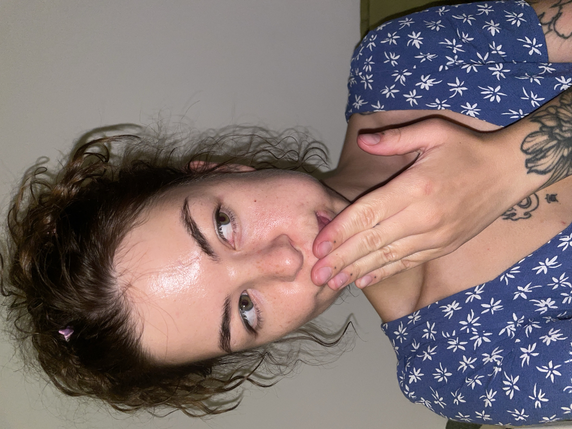 A young woman with her hair pulled up in a messy bun, wearing a blue dress with white floral patterns, covers her mouth with her hand. She has light skin, freckles, and light brown hair. A small pink clip is visible in her hair. Her left hand is positioned in front of her mouth, and her right hand is partially visible. She appears to be looking to her right. The background is a plain, light-colored wall. The scene is indoors, and the lighting suggests it is evening or dimly lit. There is no indication of the city of Boreti, Montenegro, within the image.