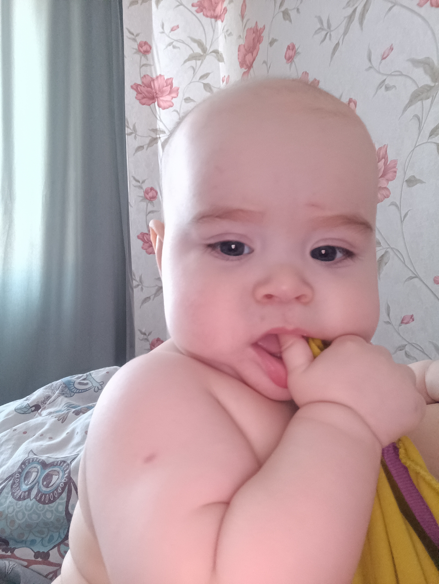 A baby is the central figure in the frame, positioned with its right hand near its mouth, holding a yellow and purple fabric. The background features a bed with a patterned owl design, a grey curtain to the left, and floral wallpaper to the right. No details suggest the location is Noviy, Russia.