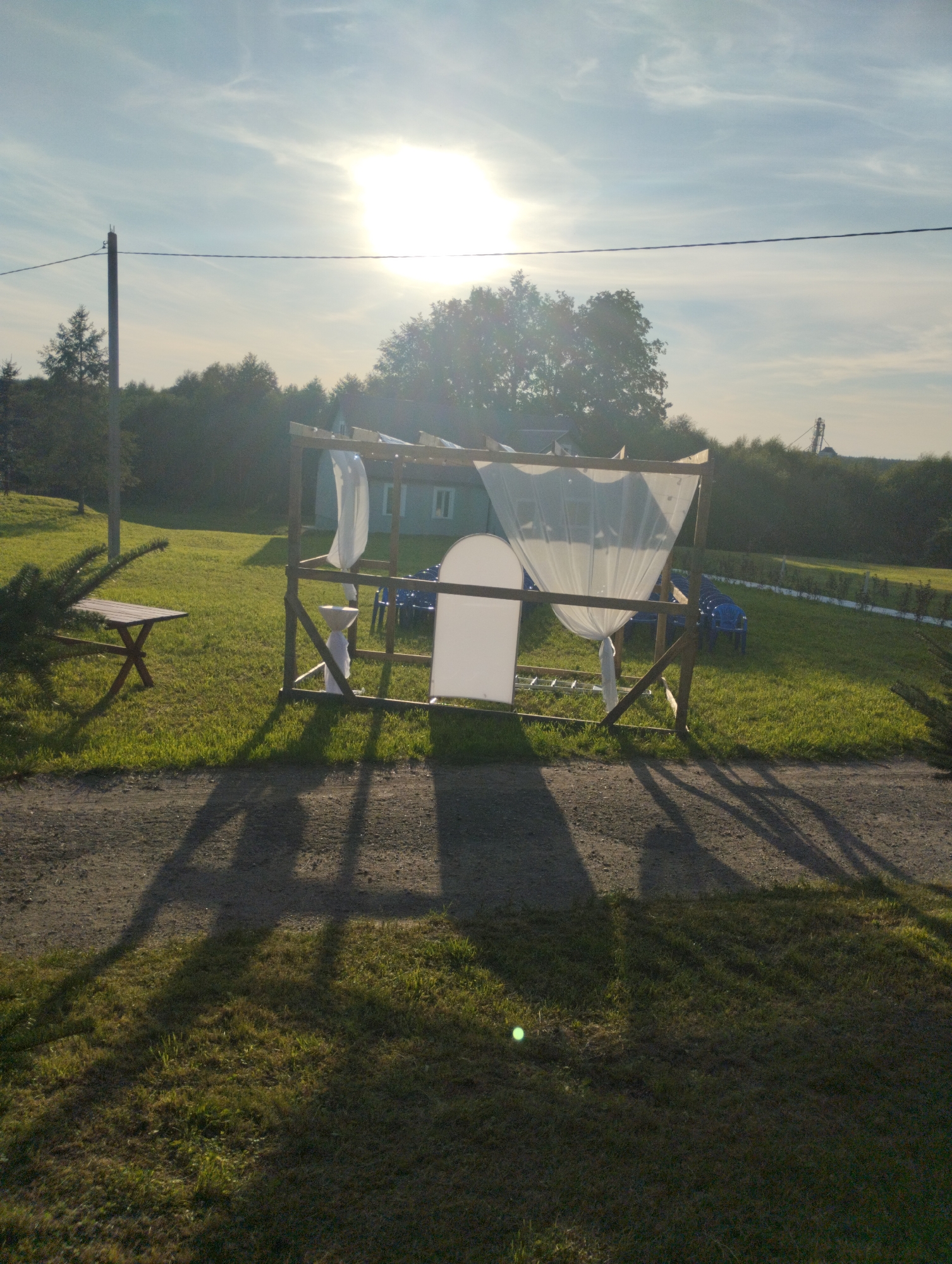 In a rural outdoor setting, likely in Вішнеўскі сельскі Савет, Belarus, a temporary structure resembling a canopy or chuppah is erected. The structure is made of wooden posts and beams, draped with white translucent fabric. Several blue plastic chairs are arranged in rows behind the canopy, suggesting a gathering or event. A wooden bench is visible to the left of the structure. The ground is covered in grass, with a gravel path in the foreground. The sun is high in the sky, casting long shadows, and trees and a building are visible in the background. The scene appears to be set during the daytime, possibly late afternoon given the lighting.