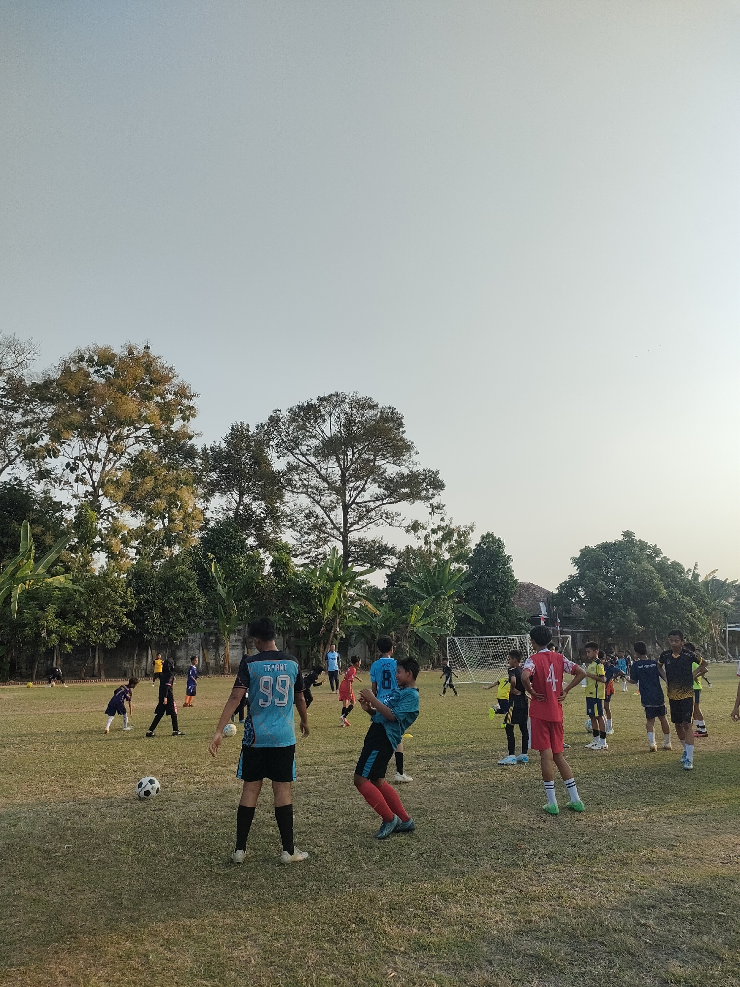 This image captures a youth soccer practice on a grassy field in Banguntapan, Indonesia. The scene features approximately 15 boys, varying in age from pre-teen to teenage, engaged in drills and informal play. Several players are wearing soccer jerseys and shorts in various colors, including blue, red, yellow, and black. One player in the foreground wears a blue jersey with the number "99" and the name "TRYANT" visible. Another player in a red jersey has the number "4" on his back. The field appears well-maintained, with a soccer goal visible in the background. Lush green trees and banana plants border the field, suggesting a tropical or subtropical climate. The sky is clear and bright, indicating daytime, likely late afternoon given the warm lighting. A few adults, possibly coaches, are present but are not the primary focus of the image. The overall atmosphere is one of energetic activity and sportsmanship.