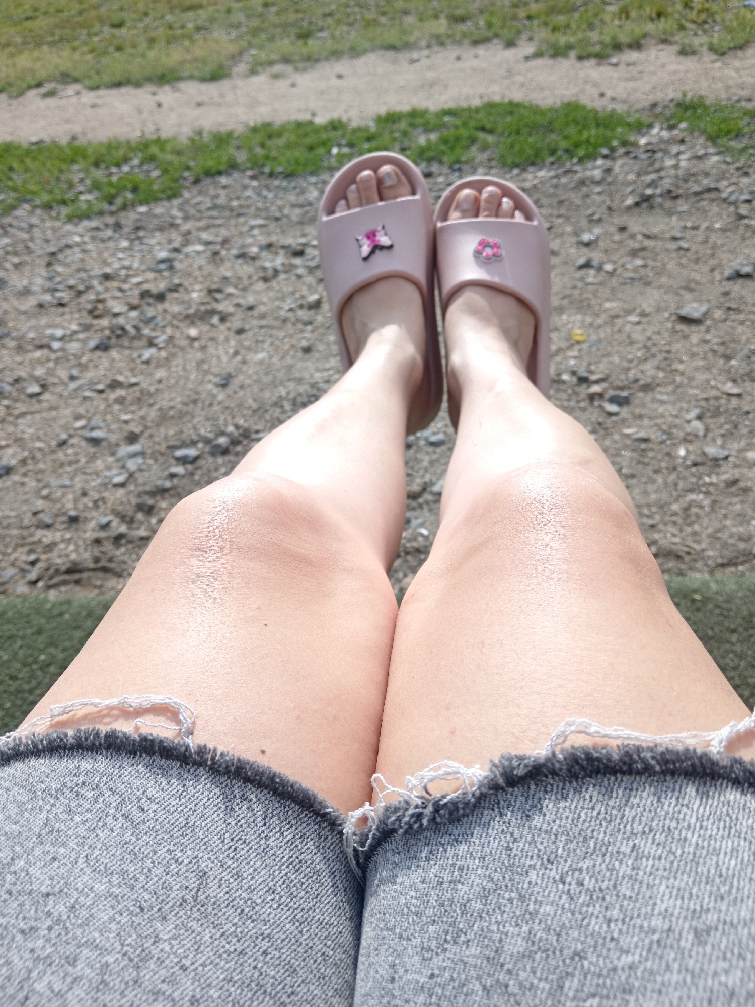 An overhead shot shows a person's legs from the knees down, resting outdoors. The person is wearing light pink slides with small flower embellishments on each. Their legs are bare, and they are wearing denim shorts with frayed edges. The ground beneath them is a mix of gravel and patchy green grass. The scene appears to be set in a park or outdoor recreational area. There is no indication of the city of Belogorsk, Russia, in the image.