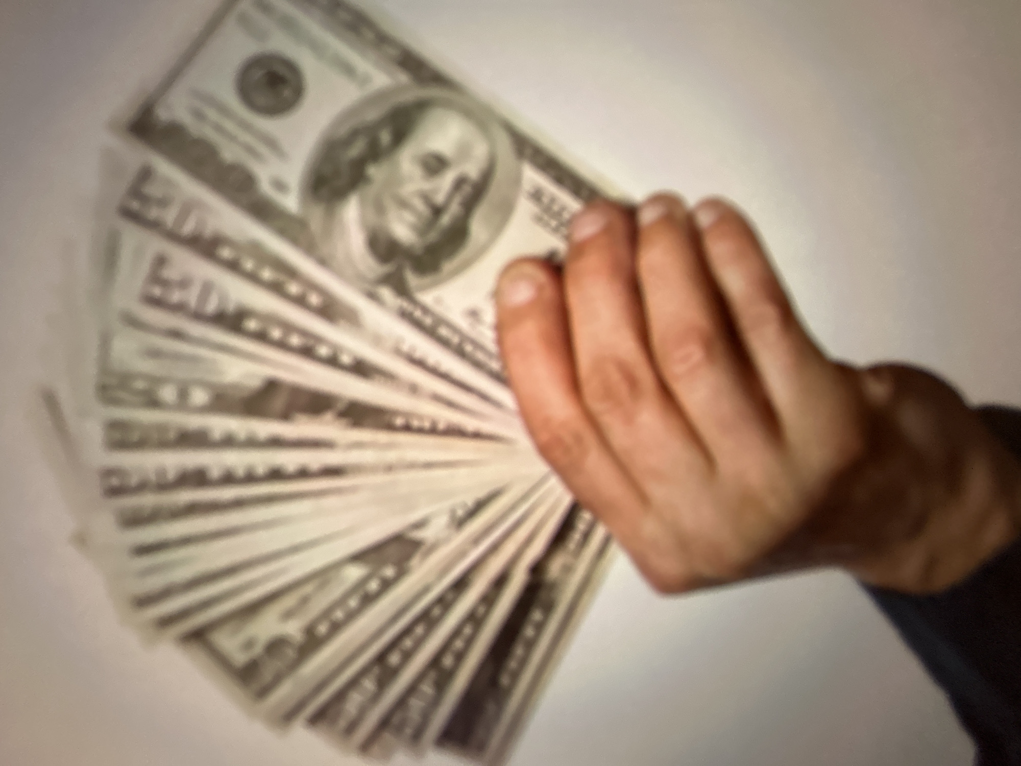 A hand is fanned out with several US dollar bills against a white background. The bills are blurry and it's difficult to determine the exact denominations, but some appear to be $100 bills due to the portrait of Benjamin Franklin visible on one of them. The hand is positioned in the lower right quadrant of the image. The background is a plain, solid white, suggesting an indoor setting. The image does not contain any information that would allow for the determination of its location in Gelendzhik, Russia, or any other specific geographic location.