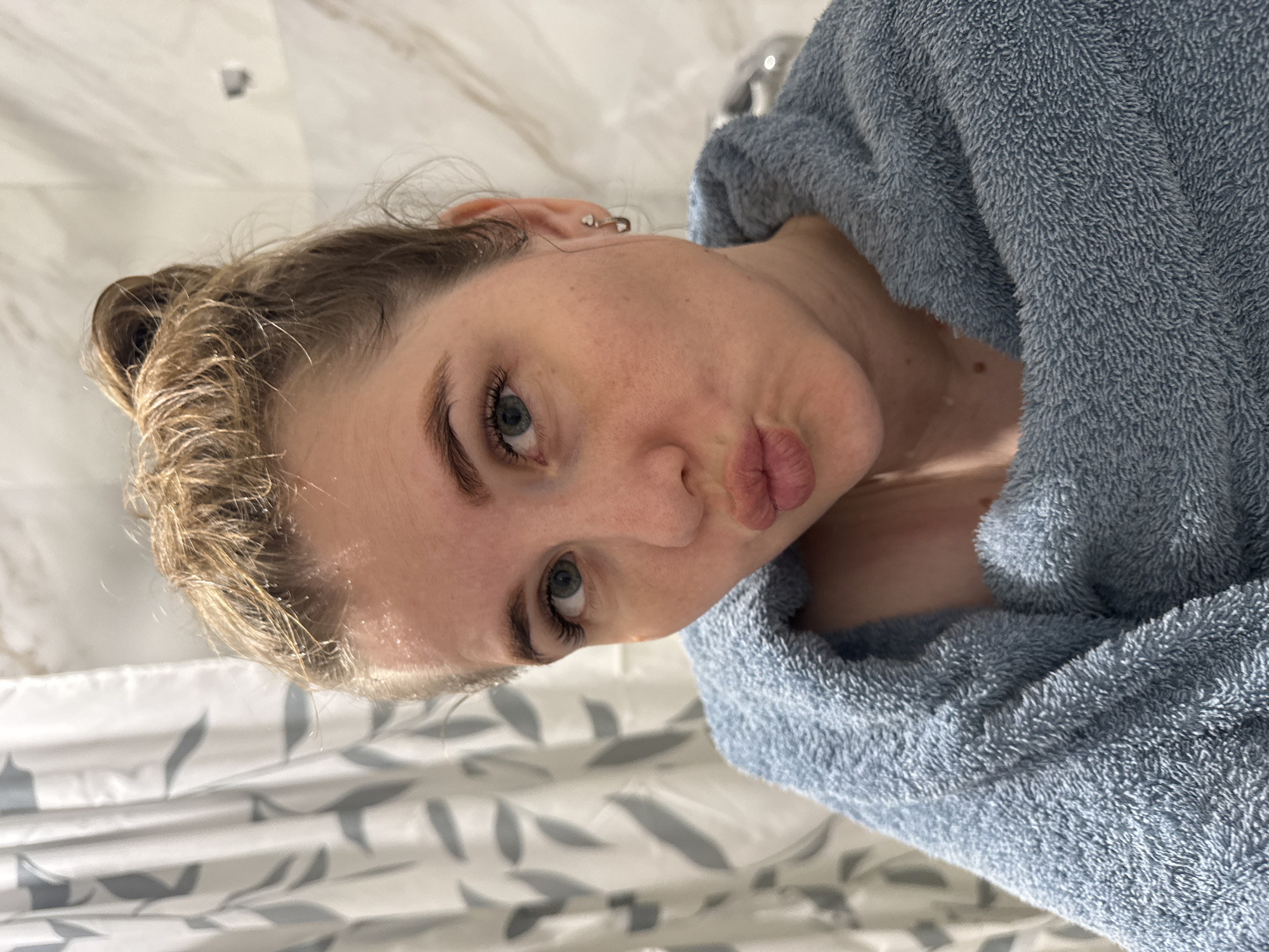 A woman wearing a blue bathrobe is posing for a selfie in a bathroom. The background shows a marble-patterned wall and a showerhead. The woman has her hair pulled back and is making a kissing face. This picture was taken in Ферма, Russia.