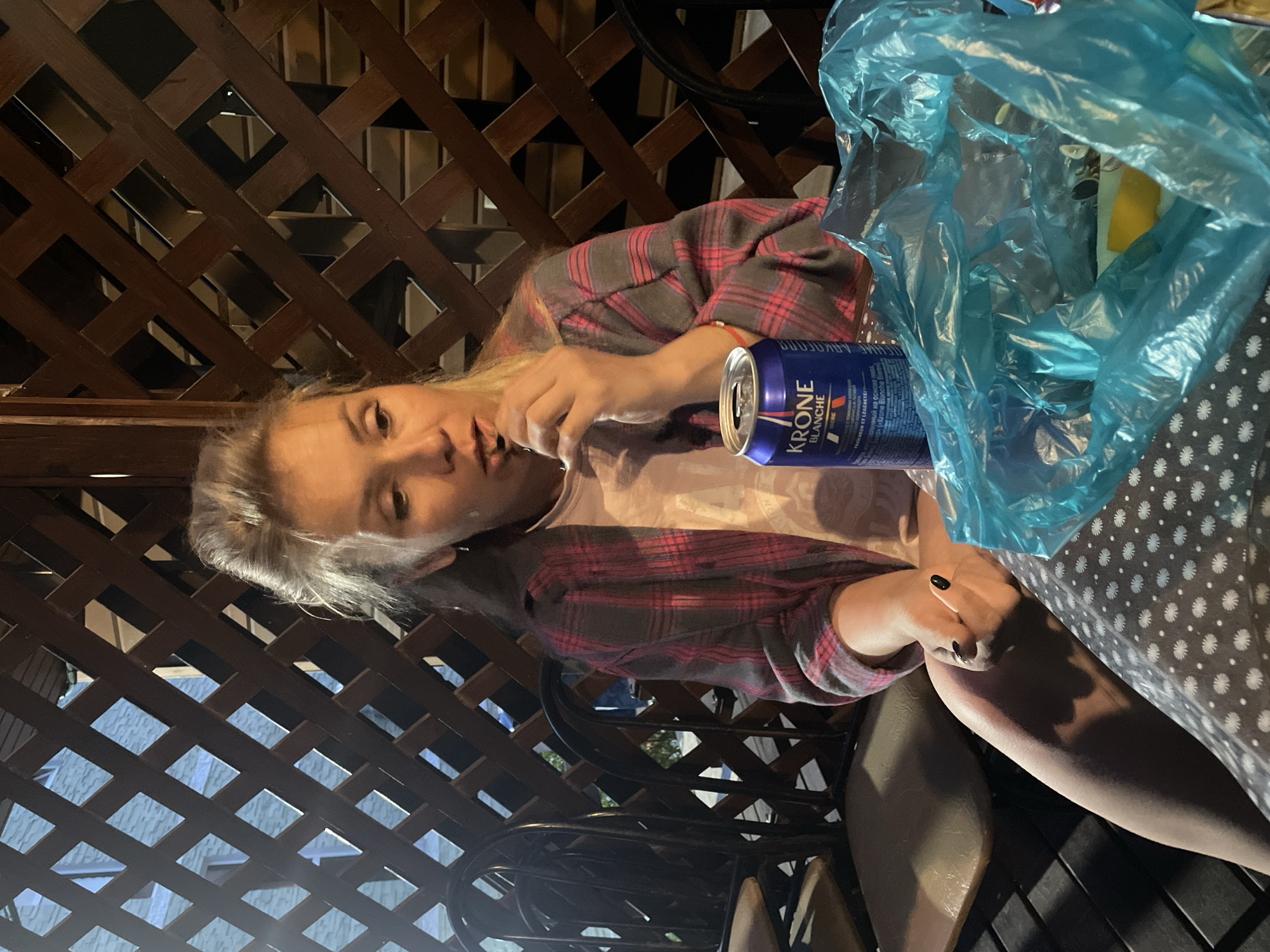 A woman with blonde hair is sitting at a table outdoors, under a wooden lattice structure. She is wearing a pink and grey plaid shirt over a light-colored t-shirt and has her right hand near her mouth, holding something dark. In front of her on the table is a blue can of "KRONE BLANCHE" beer and a bright blue plastic bag containing unseen items. The table is covered with a dark cloth with a white polka dot pattern. Behind her, dark chairs with light-colored seats are visible, and through the lattice, a blurry background of what appears to be buildings and possibly water can be seen. The lighting suggests it is evening or night.