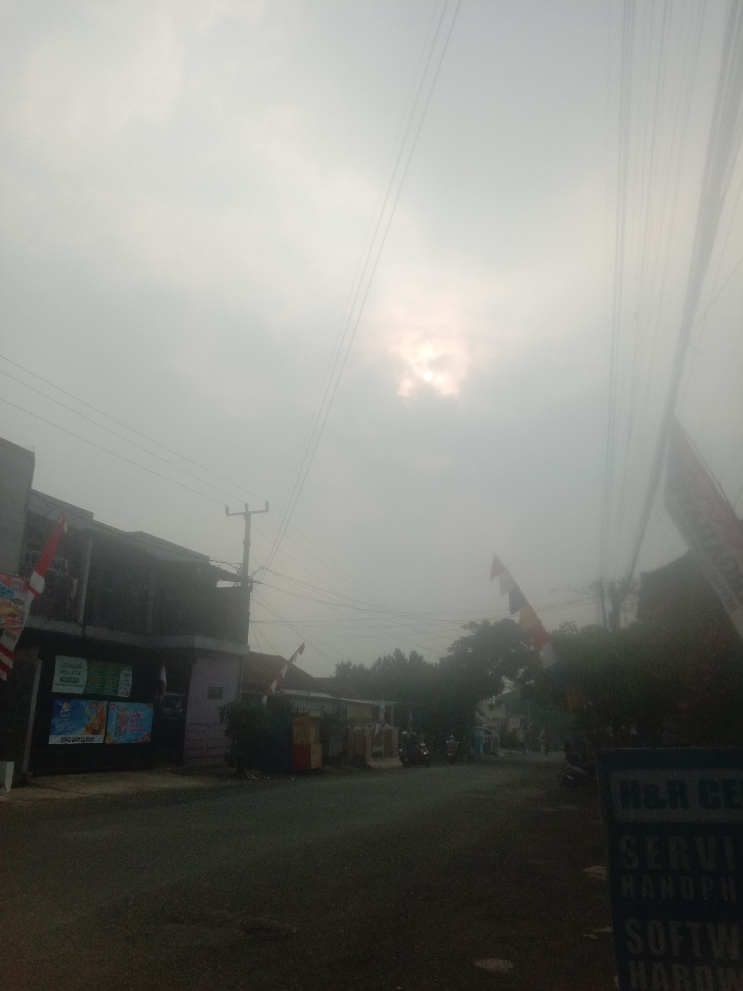 The image depicts a street scene in Bojong, Indonesia, under an overcast sky with the sun partially visible through the clouds. On the left side of the road, a two-story building with dark-colored walls is prominent. Several colorful advertisements are displayed on the lower section of this building. A red and white Indonesian flag is attached to a pole on the left side of the building. Across the street, a sign with the words "H&R CE" and "SERVI HANDPH" and "SOFTW HARDW" is visible on the right side of the road. A utility pole with electrical wires is situated near the building on the left. The road surface is asphalt and appears to be a two-lane street. In the background, several trees and other buildings can be seen, along with a few motorcycles parked or moving. The overall atmosphere is hazy, suggesting a potentially humid or polluted environment.