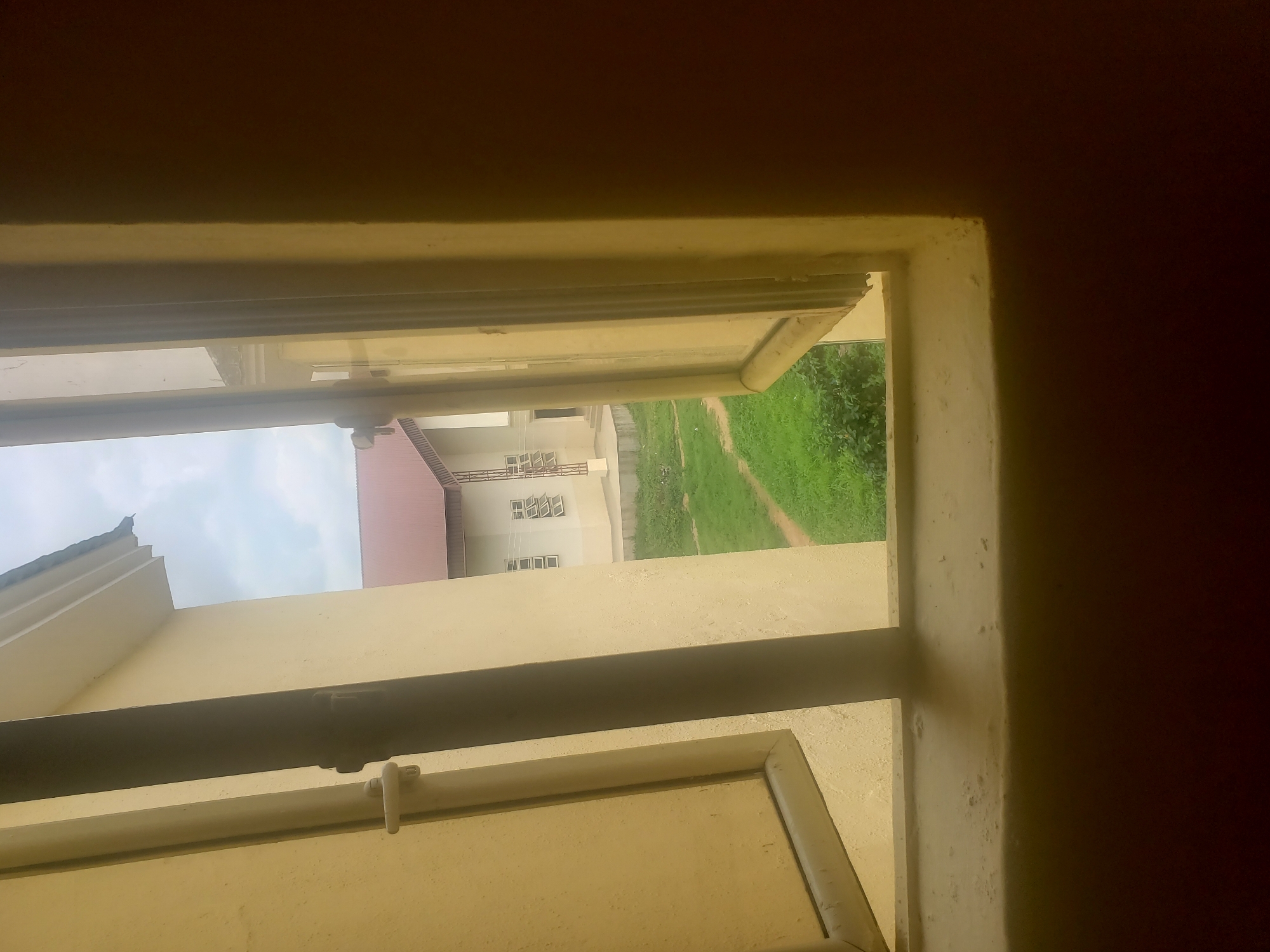 The image, captured from inside a building in Gumel, Nigeria, looks out through an open window. The window frame and sill are painted a light yellow. Through the window, a green, grassy area is visible, with a pathway cutting through it. Beyond the grass, a light-colored building with a reddish-brown roof stands under an overcast sky. The building has multiple windows, and some structural elements appear to be made of metal. The interior wall on the right side of the frame is dark brown and textured.