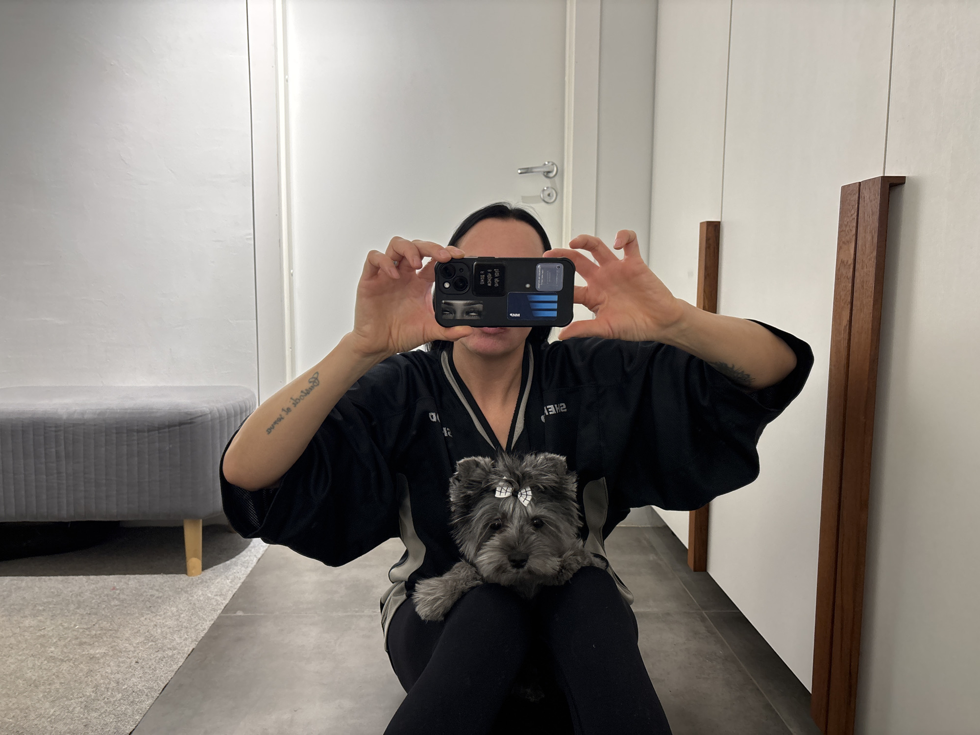 A woman sits in a room holding a cell phone up to take a picture. The cell phone has stickers on the back, including one that appears to be eyes. The woman is wearing a black top with silver accents, and black leggings. She holds a small, gray dog on her lap. The dog has a white bow in its hair. Behind the woman is a grey ottoman on the left, and a wall with several doors on the right, with wooden handles. The location is in a room in the city of Chaikovsky, Russia.
