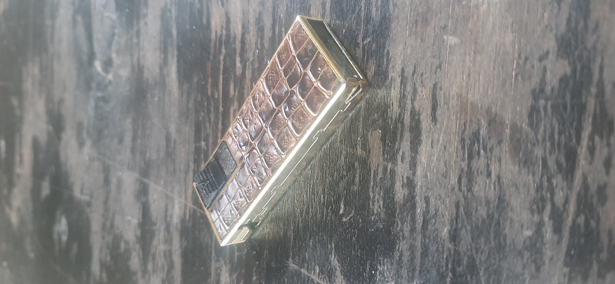 A golden lighter with a faux crocodile skin texture sits on a dark, weathered wooden surface. The lighter is rectangular with a black firing mechanism on one end. The surface it rests on is a dark wood with visible grain and wear marks. The overall lighting suggests an outdoor or dimly lit indoor setting. No specific location in Yessentukskaya, Russia, can be determined from the image.