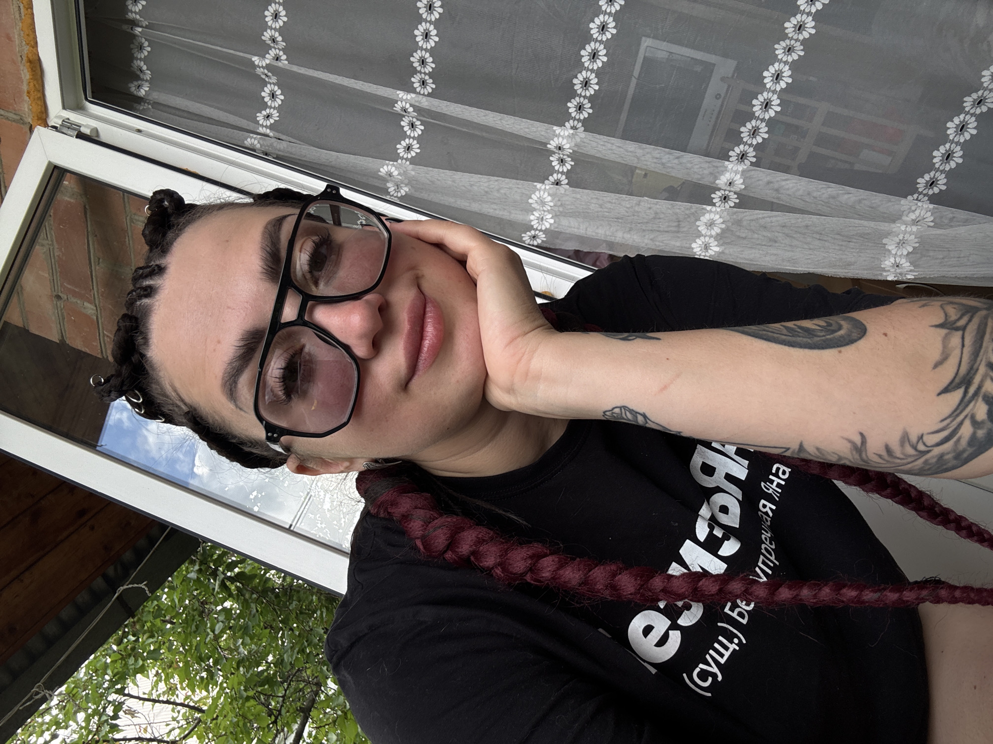 A medium shot captures a woman with dark braided hair and large black-rimmed glasses. She is wearing a black t-shirt with white lettering and has tattoos on her arm. The woman is outdoors, leaning her head on her hand, with a window and curtains visible in the background. Green foliage is also present in the upper left portion of the image. The setting appears to be a residential area.