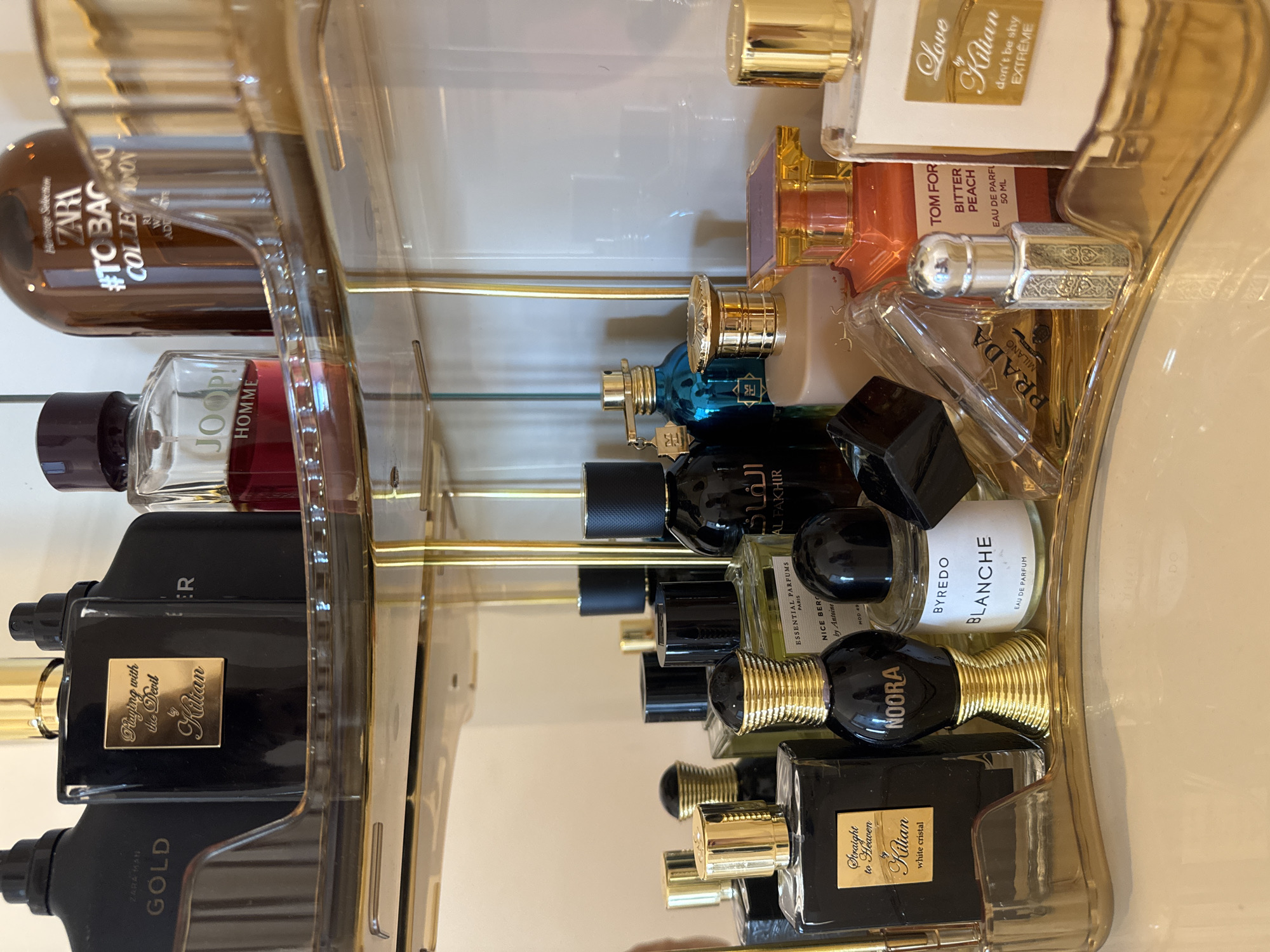 This media file is a close-up, vertical shot of a collection of perfume bottles displayed on a tiered, clear acrylic shelf with gold accents. The bottles are of various shapes, sizes, and colors, including black, brown, clear, and blue. Some bottles have ornate detailing on their caps or labels.
Visible text on the labels includes brand names like "ZARA," "JOOP!," "Kilian," "BYREDO," and "PRADA." Specific fragrance names like "Playing with the Devil," "Straight to Heaven," "GOLD," "Love," "don't be shy EXTRÊME," "TOM FORD BITTER PEACH," and "BLANCHE" are also legible.
The lighting appears to be bright and even, suggesting an indoor setting, possibly a vanity or a retail display. The focus is sharp on the perfume bottles, creating a clean and organized aesthetic. There are no people or explicit signs of a specific time of day or weather conditions. The overall impression is one of luxury and a curated collection of fragrances. The composition is tight, emphasizing the details of the bottles and their branding.