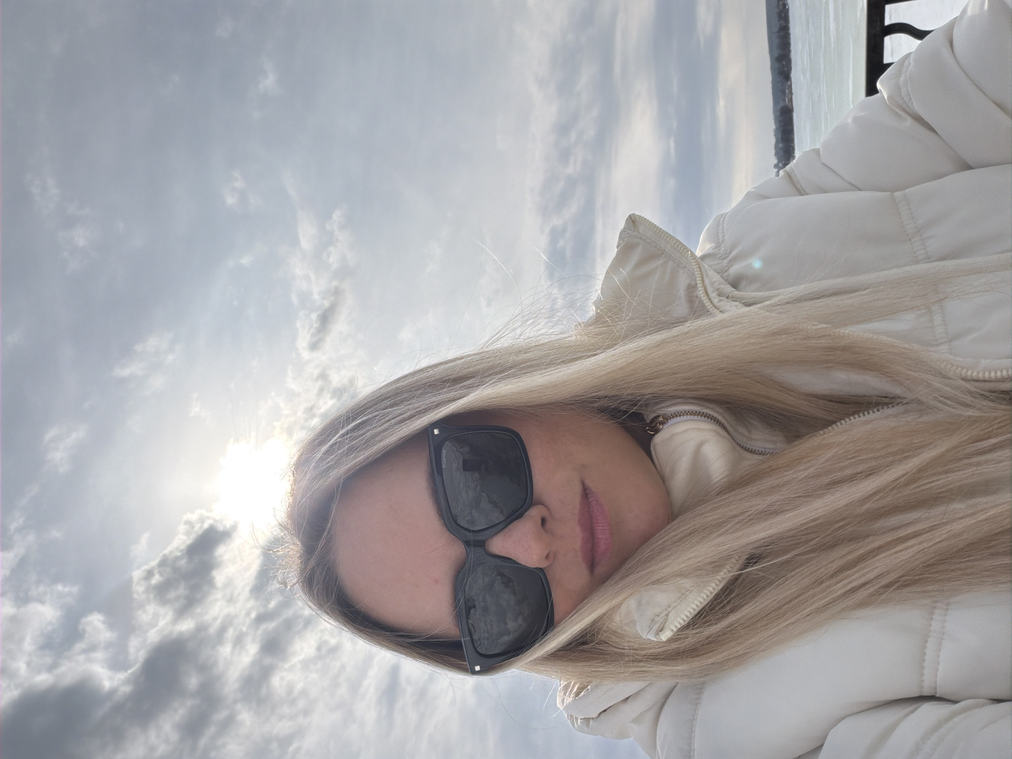 A woman with long blonde hair poses outdoors wearing sunglasses and a white puffer jacket. The background shows a cloudy sky with the sun partially obscured, and a distant view of what appears to be a body of water with some structures along the shoreline. Given the mention of Votkinsk, Russia, this could be the Votkinsk Reservoir. There's no further detail to determine the specific location.