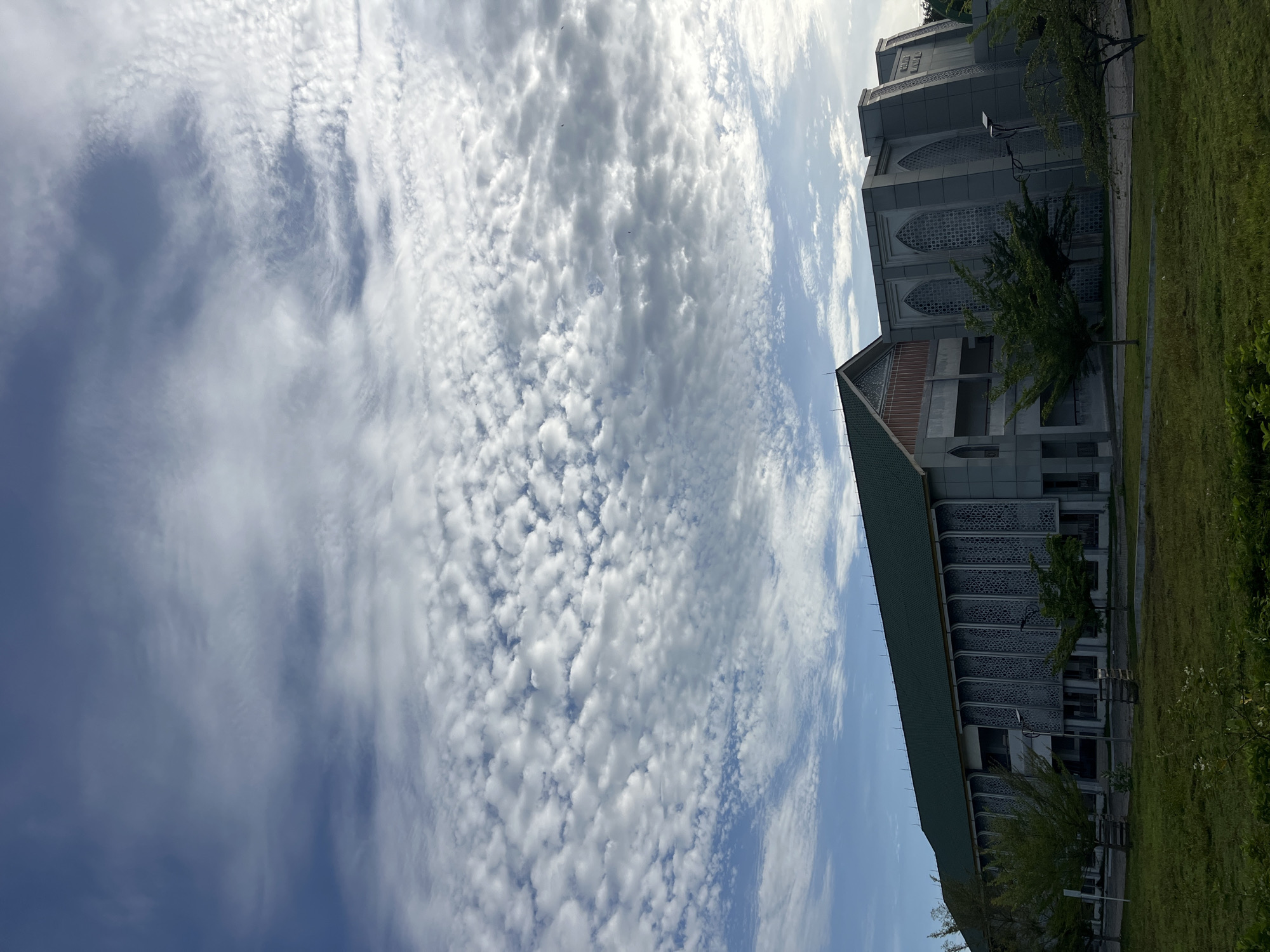 The image captures a view of the Baiturrahman Islamic Boarding School in Lhokseumawe, Indonesia. A predominantly blue sky is filled with altocumulus clouds. A building with a green roof and gray facade occupies the right side of the frame, adjacent to a grassy area. The architecture of the building features patterned latticework windows. Trees are positioned close to the building, partially obscuring its lower sections. The photograph is taken at an angle, tilting the scene to the side.