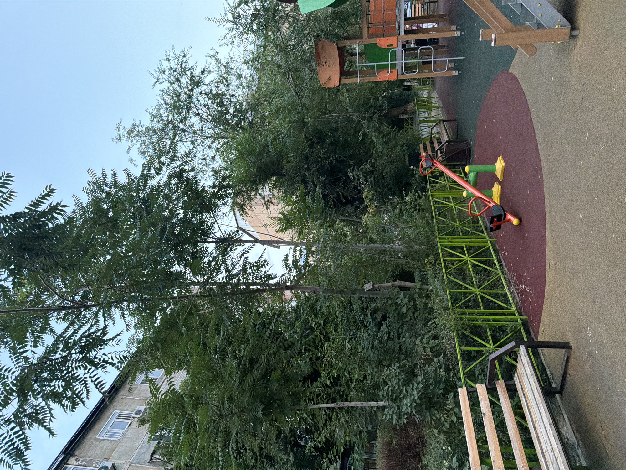 The image depicts a public playground area in the city of Shymkent, Kazakhstan. In the foreground, a wooden bench is positioned on a beige and green rubberized surface. To the right, a bright red and yellow seesaw is set up on a maroon circular section of the playground. Behind the seesaw, a portion of a larger wooden playground structure with orange and green elements is visible. A bright green metal fence with a geometric pattern runs across the middle ground, separating the playground from a densely vegetated area. Beyond the fence, several tall trees and the side of an apartment building are visible. The sky above is clear and blue, suggesting daytime. The overall scene conveys a typical urban park environment.