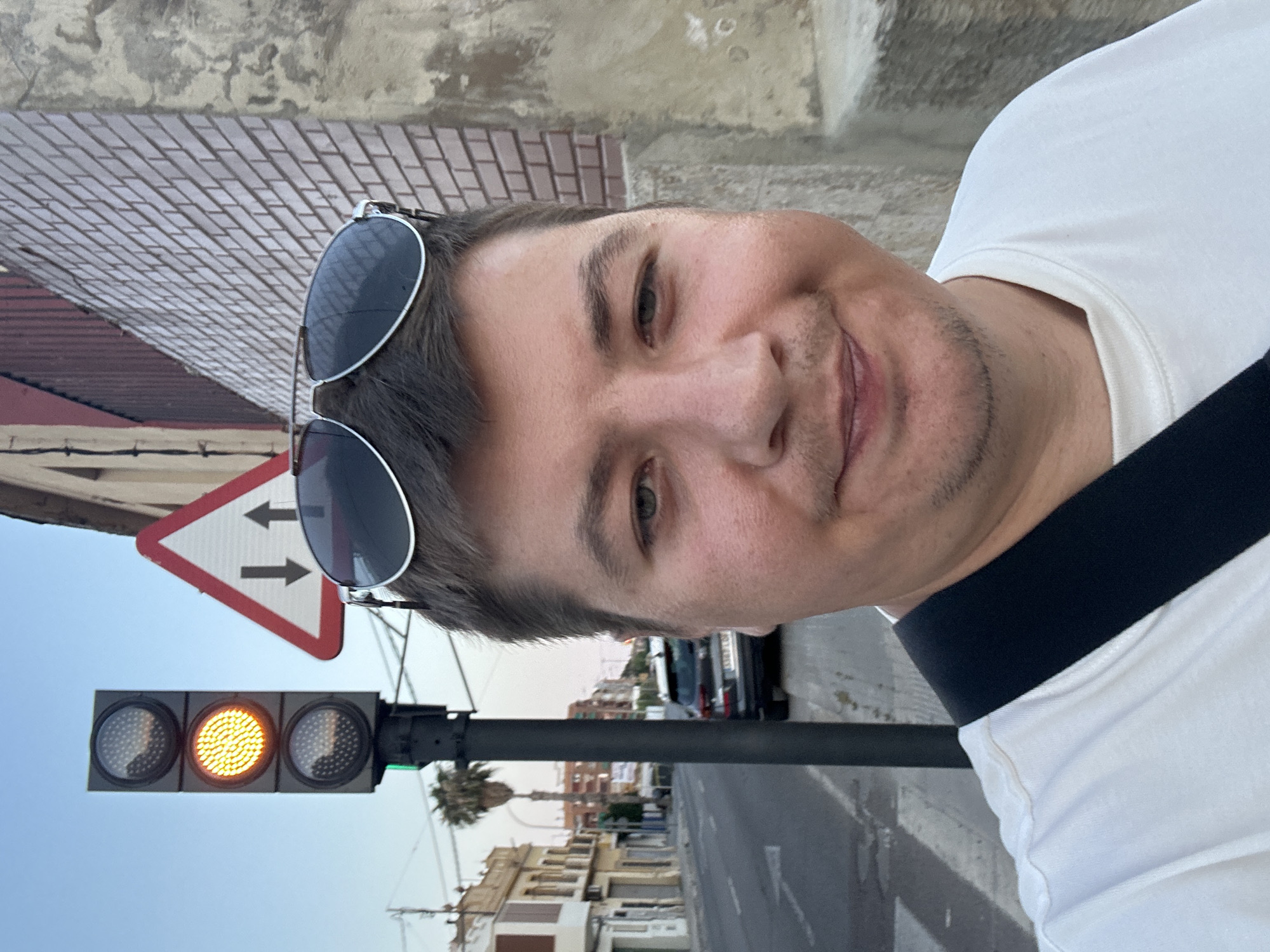 The image shows a man with sunglasses on his head, taking a selfie on a street in Benimàmet-beniferri, Spain. A traffic light is visible on the left, with the yellow light illuminated. A warning sign with two arrows pointing in opposite directions is also present. The background includes buildings and a street with a parked car. The man is wearing a white t-shirt and a black strap across his shoulder. The overall scene appears to be a daytime shot under a clear sky.