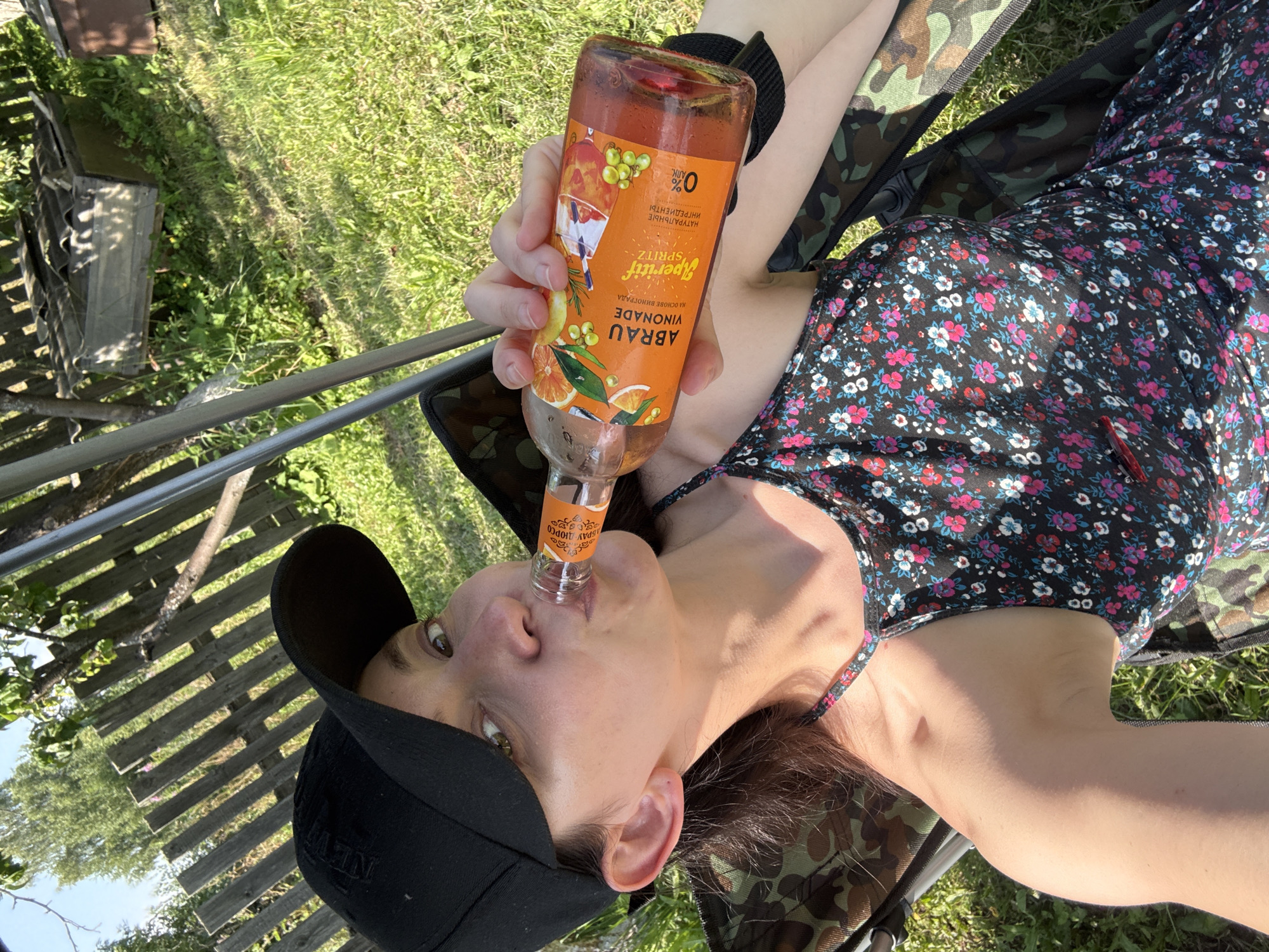 A woman is sitting in a camouflage camping chair outdoors, in what appears to be a backyard in Zasechnyi, Russia. She is wearing a black baseball cap and a black floral-print dress. The woman is holding a bottle of "Abrau Limonade Spritz" and drinking from it. The background consists of a wooden fence, green grass, trees, and some rustic structures. The lighting suggests it is a sunny day.