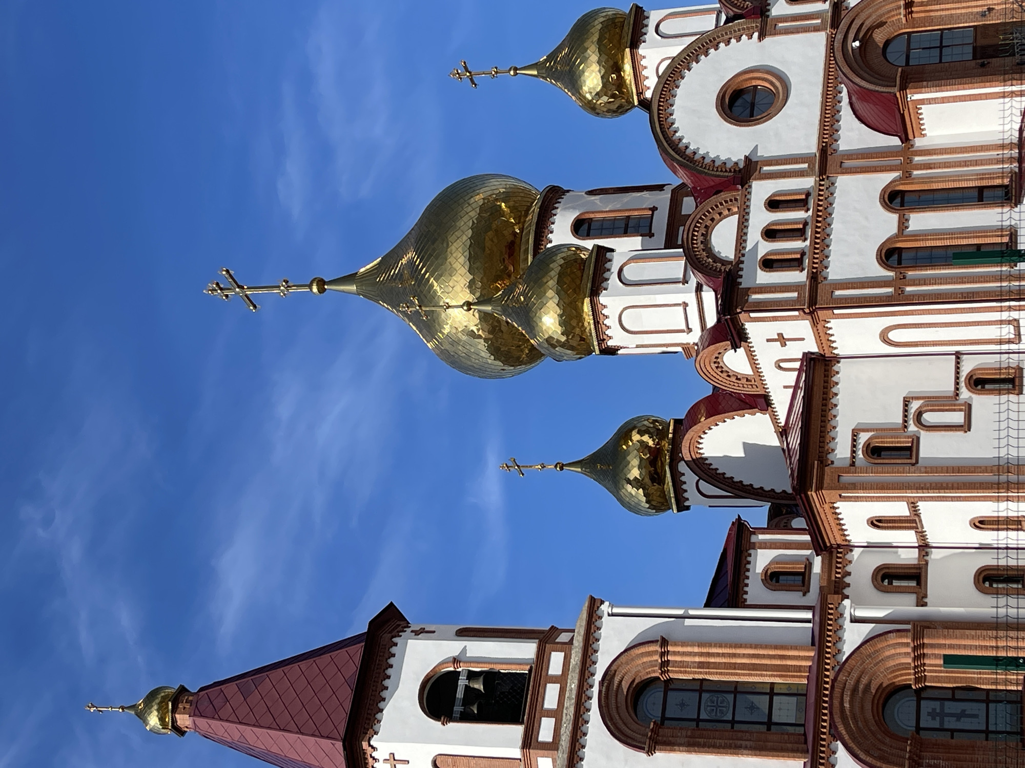 The image depicts a portion of the Krasnoyarsk Saints Peter and Fevronia Church against a bright blue sky. The architectural details consist of white walls with red and brown brick accents. Several gold-domed onion structures are topped with Orthodox crosses. A bell tower with a red tile roof is also visible. The photograph is taken from a low angle, emphasizing the height and ornate features of the church.