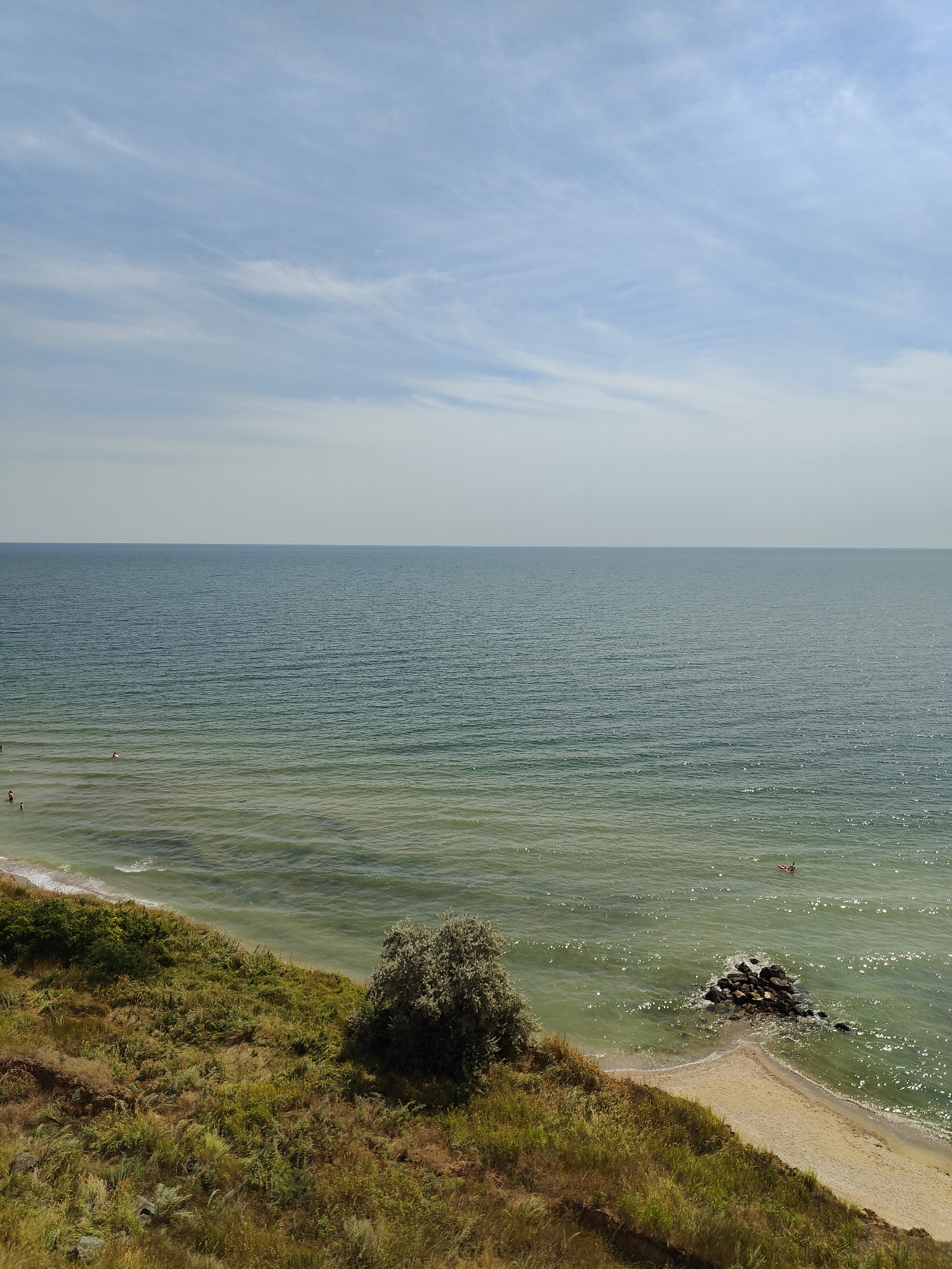 The image depicts a scenic view of the Black Sea coast near Sanzhiika, Ukraine. The scene is dominated by the vast expanse of the sea, which occupies the majority of the frame. The water is a muted green-blue, with gentle waves creating a textured surface that shimmers in the sunlight. In the distance, the horizon is clearly defined, separating the sea from a pale blue sky streaked with wispy white clouds.
In the foreground, a grassy cliff overlooks the sea. The vegetation is a mix of green and dry grasses, with a few small bushes and trees scattered across the slope. A small sandy beach is visible at the base of the cliff, leading to the water. A cluster of dark rocks protrudes from the sea near the shore.
There are a few individuals in the water, suggesting recreational activity. One person appears to be swimming or floating on an inflatable toy, while others are closer to the shore, wading or swimming. The overall atmosphere is one of natural tranquility and a pleasant day at the seaside.