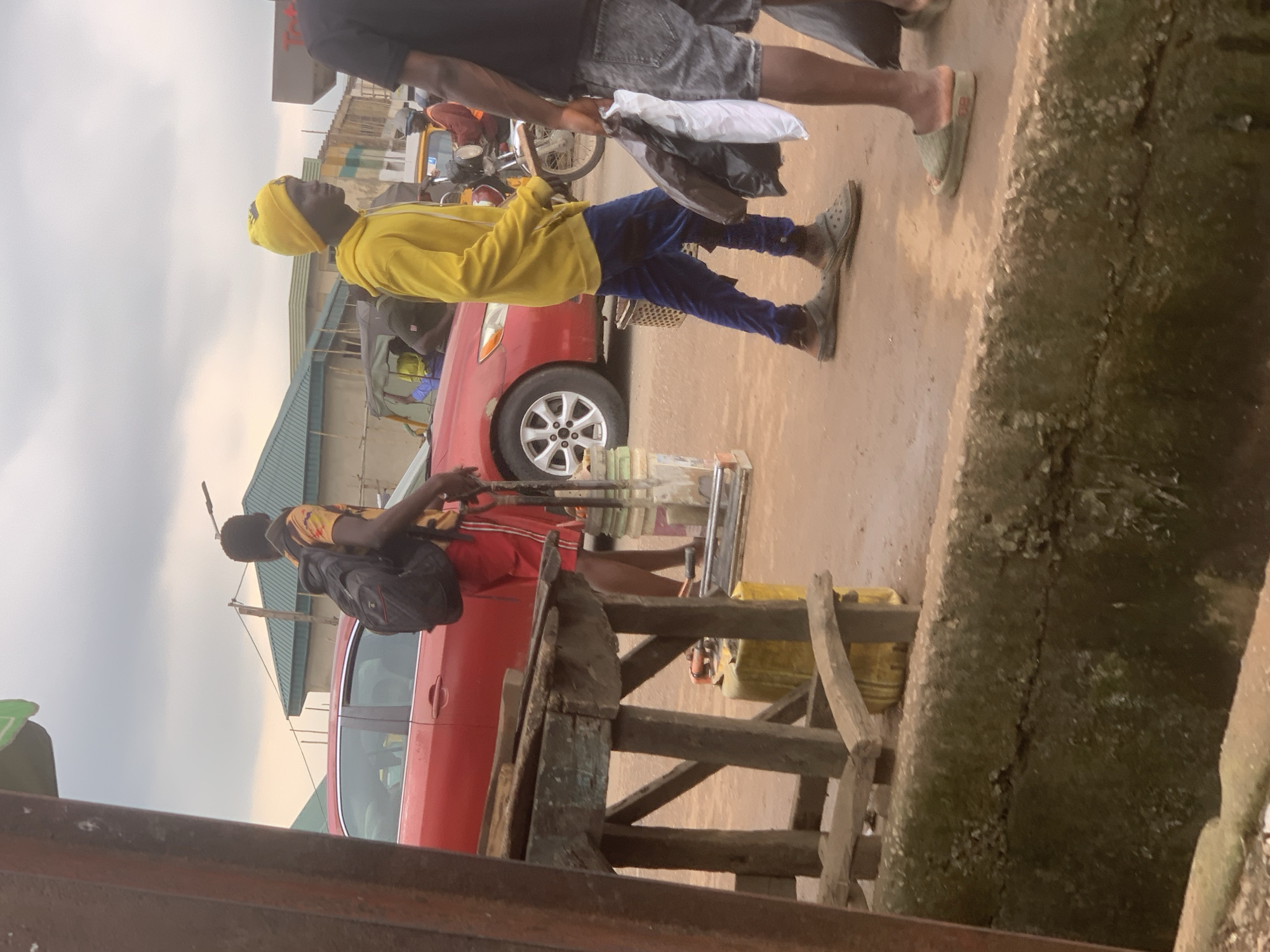 The image shows a street scene in Shagamu, Nigeria, with a focus on pedestrians and vehicles. In the foreground, a man wearing a yellow hoodie and blue pants, topped with a yellow beanie, walks towards the right. He is carrying two plastic bags, one black and one white. To his left, another person with a black backpack and red shorts is walking away from the camera. Behind them, a red sedan is parked or moving slowly. Further back, a motorcycle and a tricycle are visible, indicating traffic. The environment appears to be an urban setting with buildings and a cloudy sky. There is a wooden structure in the left foreground and a concrete ditch running along the bottom of the image.