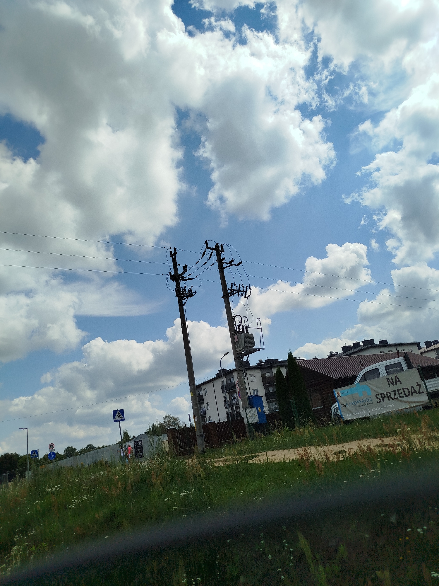 The image captures a street scene in Wieliszew, Poland, on a bright, cloudy day. Dominating the upper portion of the frame are vast expanses of sky filled with fluffy white clouds against a vibrant blue backdrop. In the middle ground, a rural road runs horizontally, bordered by lush green grass and wildflowers. A concrete fence runs along the side of the road, separating it from the background. Beyond the fence, several multi-story buildings, typical of a suburban or town setting, are visible. Adjacent to one of the buildings is a dark wooden structure. A white van is parked nearby, with a large banner on its side that reads "NA SPRZEDAŻ" (For Sale) in Polish, along with company information. Two power poles with electrical equipment are prominently positioned in the mid-ground, with wires stretching across the frame. Various street signs are also present, including pedestrian crossing signs and a circular traffic sign. A few individuals are casually walking near the fence in the middle ground. The overall impression is a typical day in a Polish town, with a mix of residential buildings, infrastructure, and natural elements.