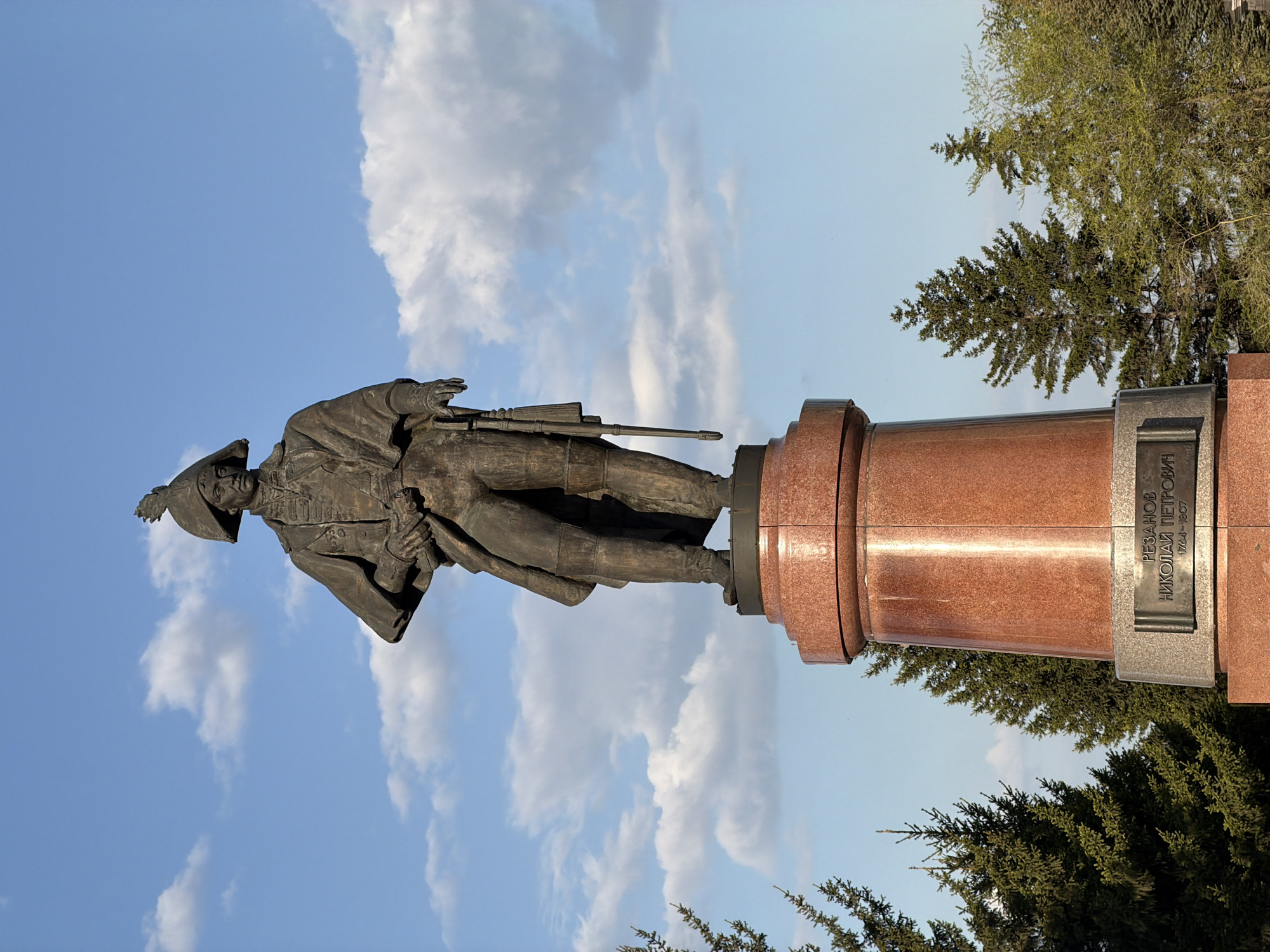 The image shows a monument of Nikolai Petrovich Rezanov in Krasnoyarsk, Russia, seen against a backdrop of a blue sky with scattered clouds and nearby trees. The bronze statue depicts Rezanov in historical clothing with a rifle, standing atop a red granite pedestal with an inscription in Russian.