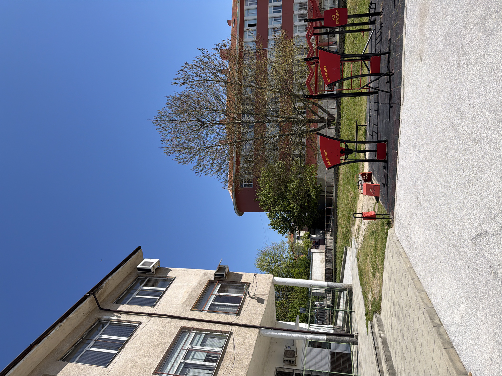 The image depicts an outdoor scene likely located in an urban environment, possibly Panagyurishte, Bulgaria. The photo shows a residential building with visible air conditioning units on its exterior wall, positioned to the left side of the frame. The building appears to have multiple windows, some with protective metal grids. Above and behind this structure is another building, taller and painted in a red color. In front of the red building, there is a tree with budding leaves and a public exercise area with red and black fitness equipment. A concrete walkway is present on the right side of the image. The sky is clear and blue, indicating fair weather.