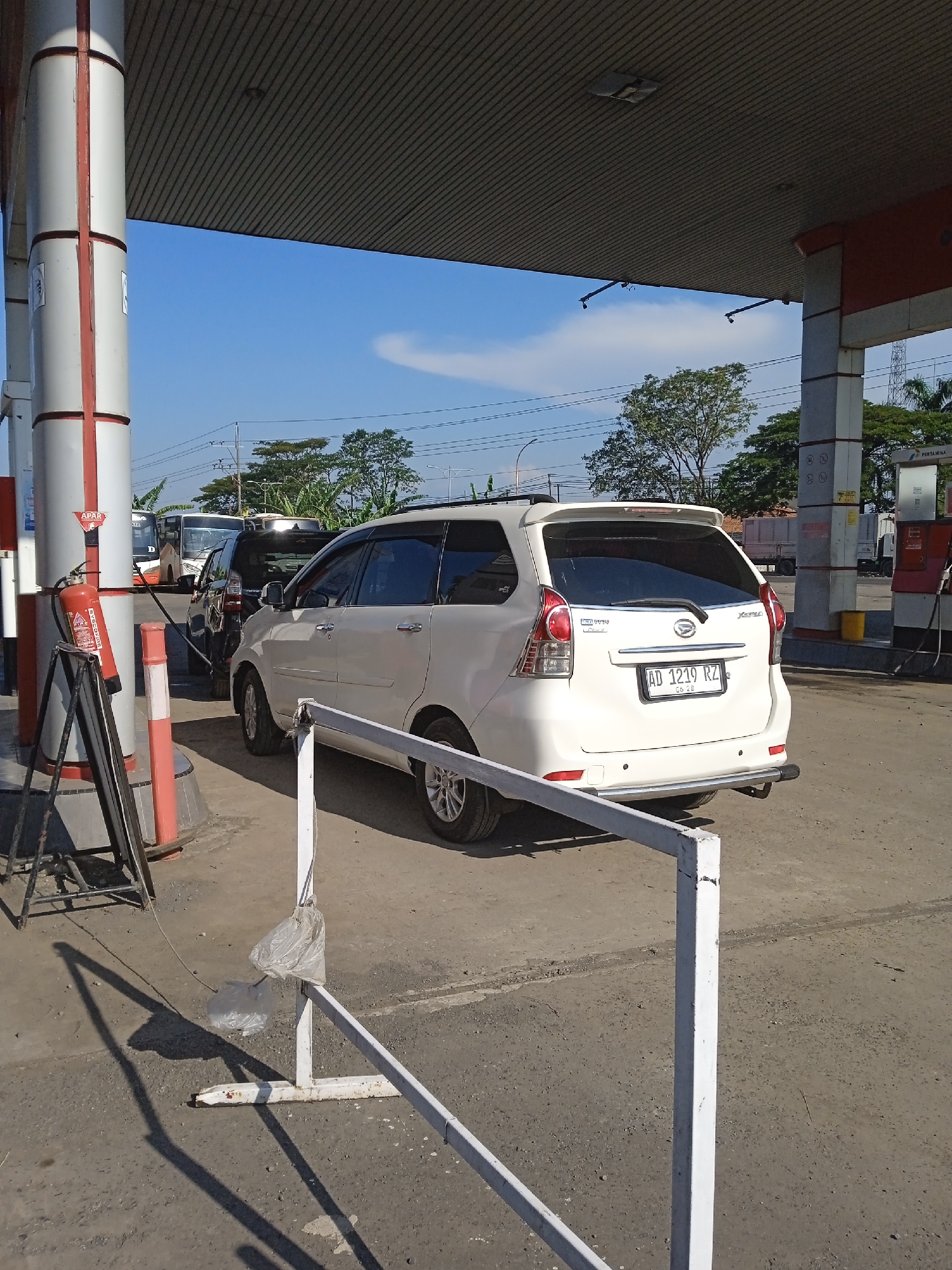 The image captures a scene at a Pertamina gas station in Brangsong, Indonesia, under a bright, partly cloudy sky. A white Daihatsu Xenia with license plate "AD 1219 RZ" is parked at one of the gas pumps. Other vehicles, including a dark-colored car and a bus, are waiting in line behind the Xenia. A red fire extinguisher stands near the gas pump. A white metal barrier is in the foreground. Trees and utility poles are visible in the background.