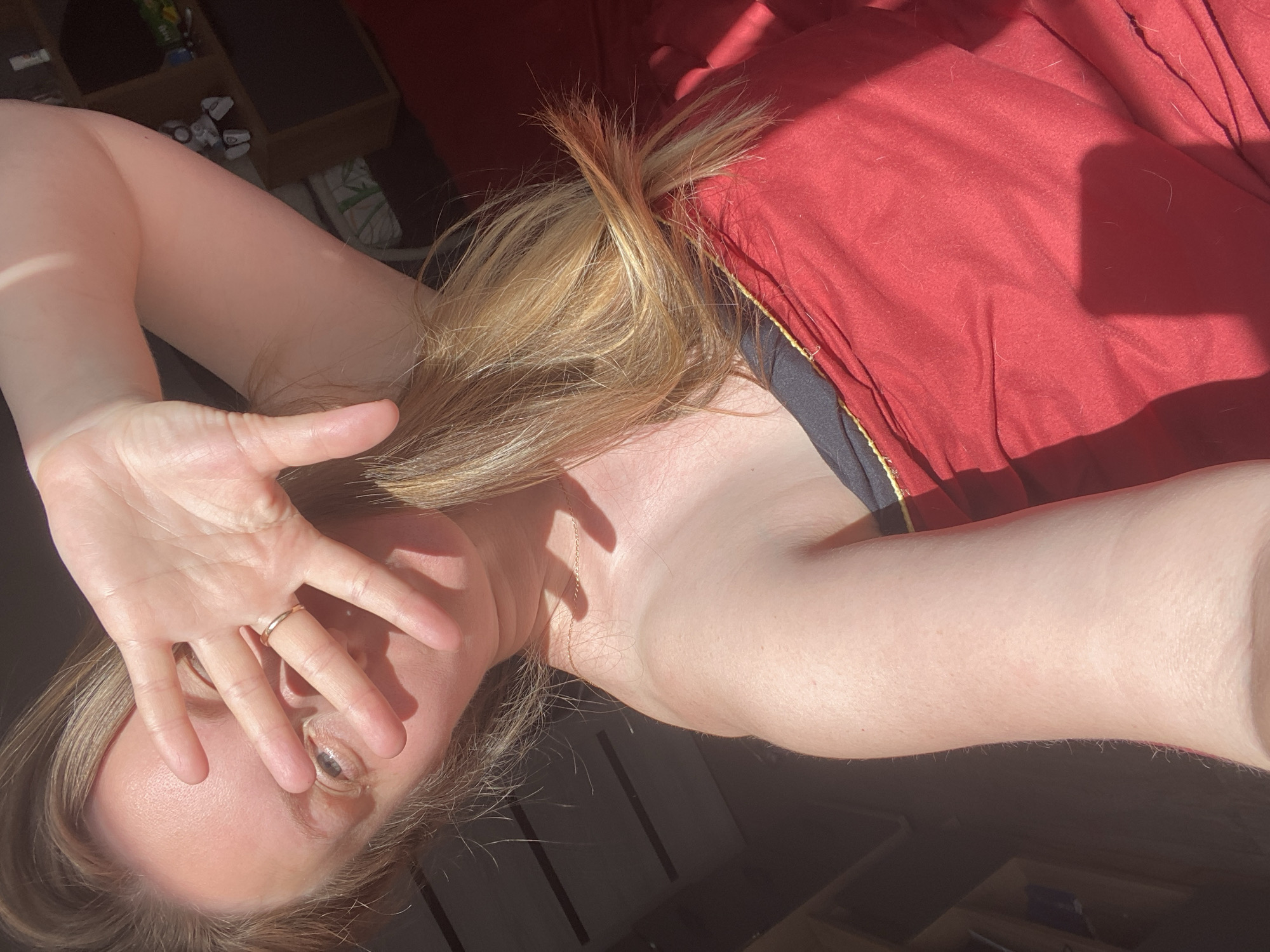 A woman with fair skin and blonde hair is lying down. She is wearing a red garment, possibly a robe or dress, with black and gold trim. Her right hand is raised, fingers spread, partially obscuring her face. A gold ring is visible on her ring finger. The scene is lit with strong sunlight, creating shadows. The location is unknown and no city or country can be determined from the image.
