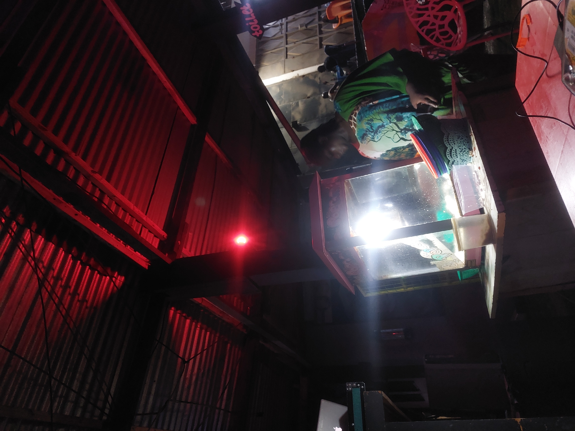The image shows the interior of a dimly lit space in Dar es-Salaam, Tanzania. Corrugated metal walls are illuminated by red lighting. A person wearing a green and blue patterned top is bending over what appears to be a display case with a bright internal light. Various objects are visible on a red countertop, including a red basket and what looks like cleaning supplies. A sign with the word "toilet" is partially visible in the upper portion of the image. A laptop with an Apple logo is positioned in the foreground, partially obscured by other objects.