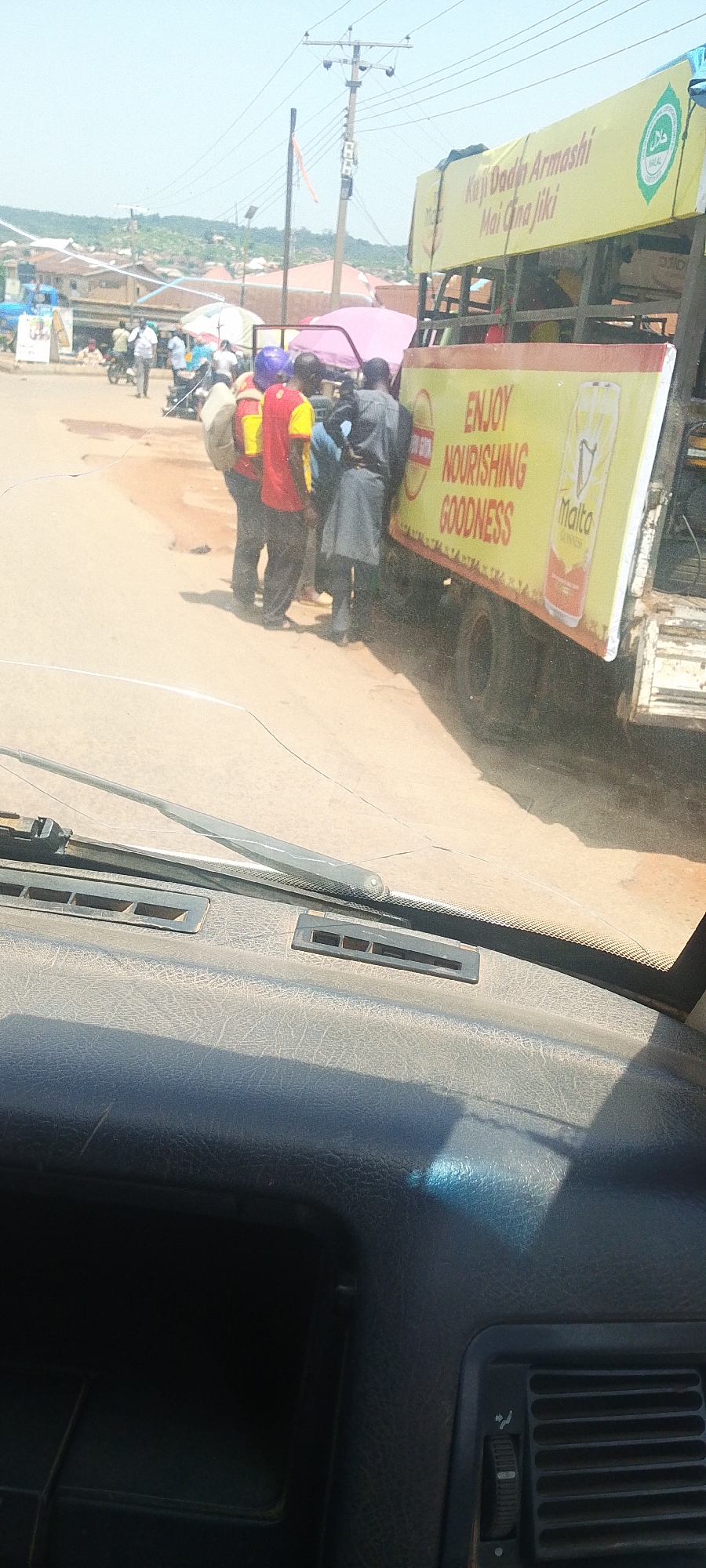 The image captures a street scene in Jos, Nigeria, with a truck prominently displayed. The truck has a large yellow banner with red lettering that reads "ENJOY NOURISHING GOODNESS" and displays an image of a Malta Guinness bottle. Another banner on the truck has text in what appears to be Hausa, including "Kuji Dadin Armashi Mai Gna Jiki" and a Halal certification mark. Several people are gathered around the truck, some wearing distinctive red and yellow vests, suggesting they might be vendors or workers associated with the truck. In the background, there are other vehicles, market stalls, and buildings, typical of a bustling urban environment. The road surface is paved, and power lines are visible overhead, indicating the presence of electricity infrastructure. The overall atmosphere suggests a commercial or distribution activity taking place on a sunny day.
