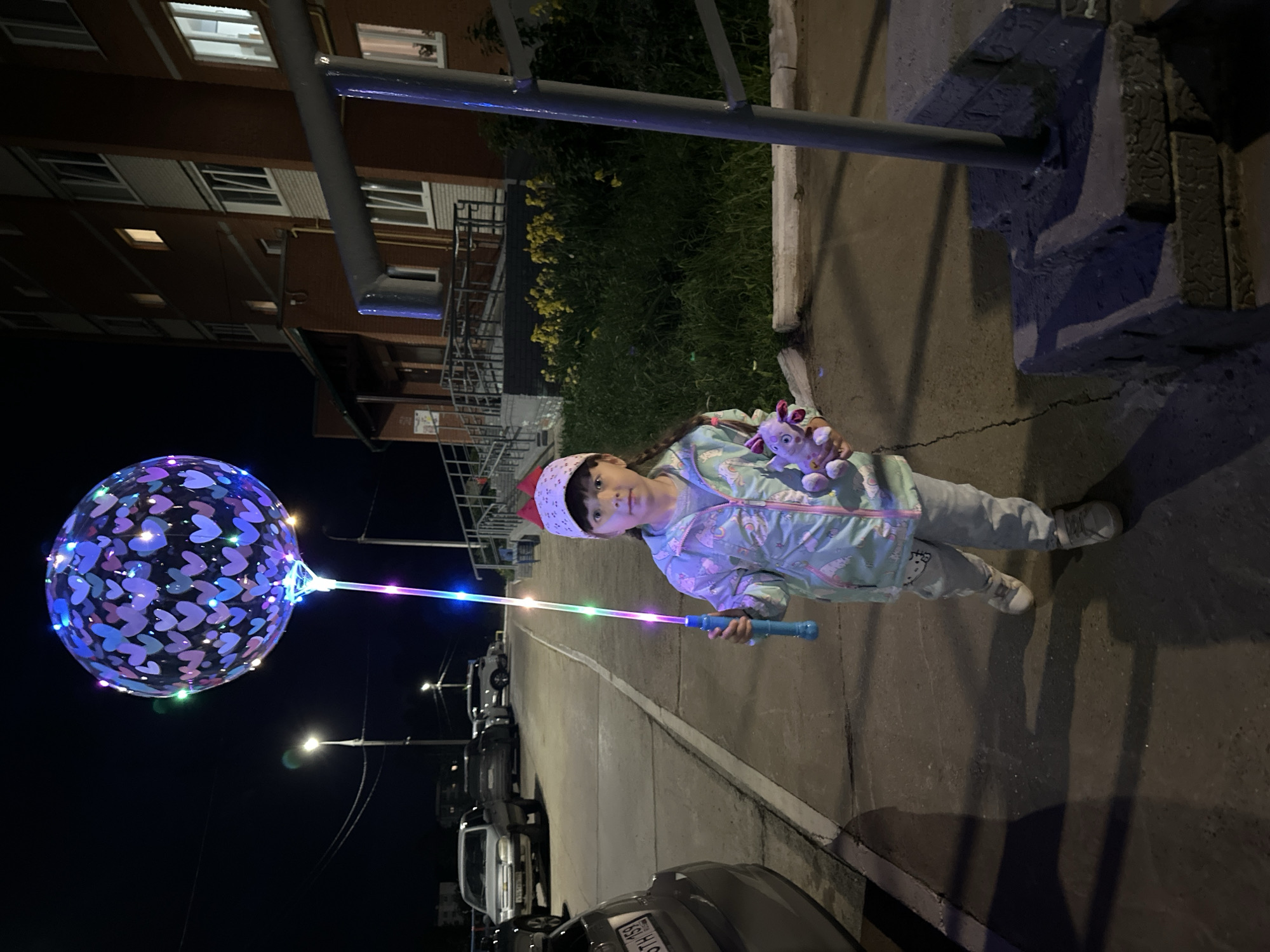 A young girl stands on a paved walkway in front of a brick apartment building at night. She is holding a transparent, heart-patterned balloon filled with lights and a light-up stick. She is wearing a light-colored jacket with colorful patterns, light-colored pants, and a red and white patterned hat. She is also holding a small stuffed toy. The walkway is cracked, and shadows are cast on it from the surrounding environment. Cars are parked in the background, and streetlights illuminate the area. The scene appears to be in Chaikovsky, Russia, given the apartment building architecture and license plates on the cars.
