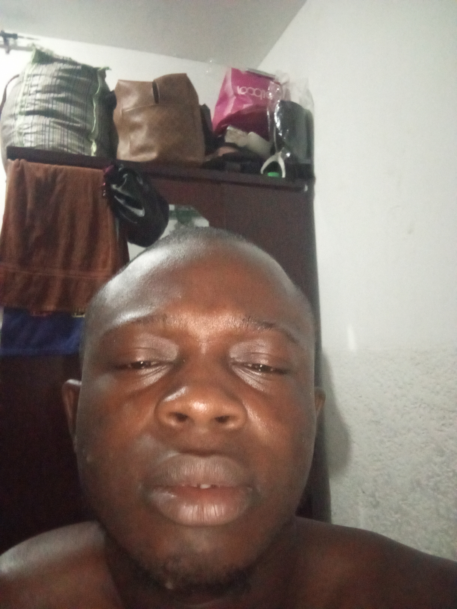 A close-up self-portrait of a dark-skinned man with a shaved head. His eyes are nearly closed, and his expression is neutral. Behind him is a dark brown armoire. On top of the armoire are several items, including a gray patterned duffel bag, a brown tote bag, a pink shopping bag, and a black item. A brown towel is hanging on the side of the armoire, with a blue item underneath. The background wall is plain white. There is no indication of the location being Abuja, Nigeria.