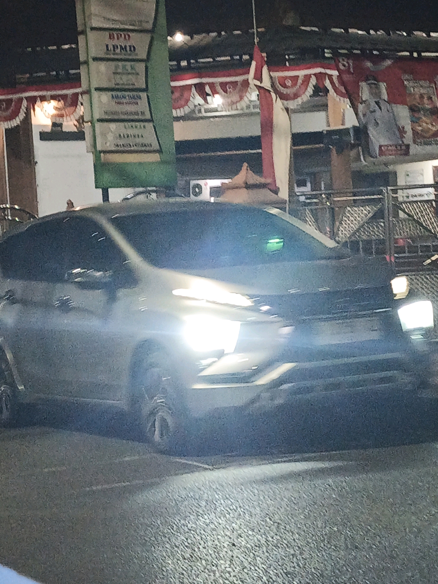 In Ketegan, Indonesia, a dark-colored, four-door SUV is parked on a paved surface, possibly a road or parking lot, at night. The SUV's headlights are illuminated, casting a bright glow that obscures some details of the vehicle and its surroundings. In the background, a tall, green signpost displays several panels with text, appearing to be official or informational. Red and white bunting is draped across the top of a structure behind the signpost, and a flag, partially visible, hangs from a pole. The overall scene suggests an outdoor setting, possibly a public area or a business establishment in Ketegan.