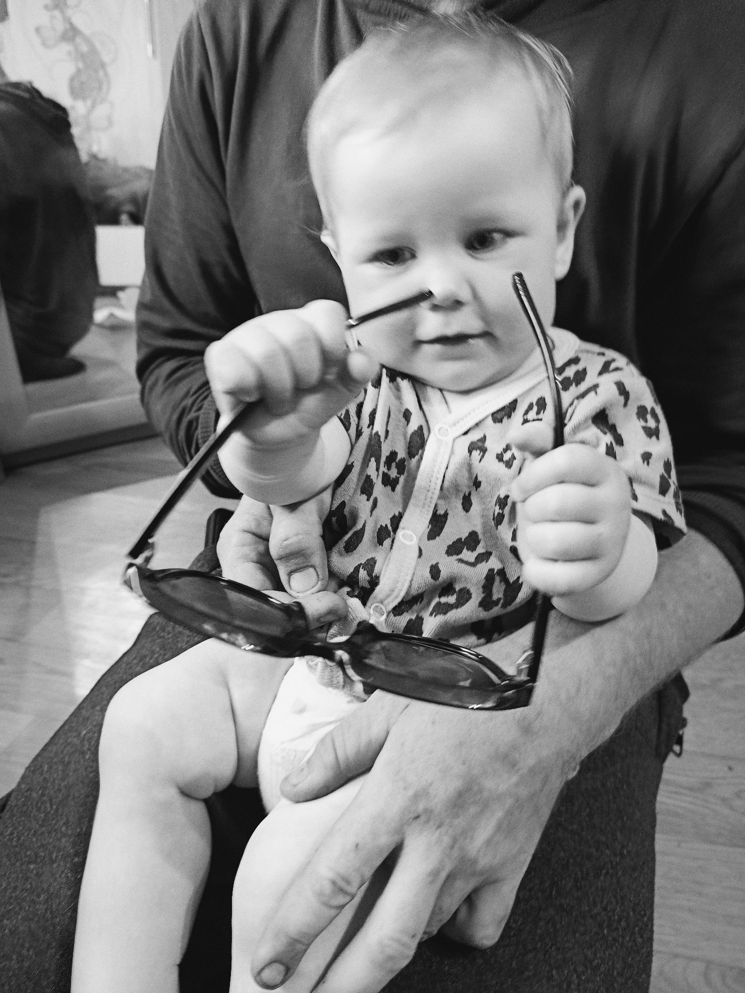 A black and white, eye-level, medium shot shows a baby sitting on someone's lap. The baby is wearing a leopard-print onesie and a diaper. The baby is holding a pair of dark-rimmed sunglasses, bringing them up to their face. The baby has blonde hair and is looking slightly to the right. The person holding the baby is wearing a dark long-sleeved shirt. The background is out of focus and appears to be an indoor setting with wooden flooring.
