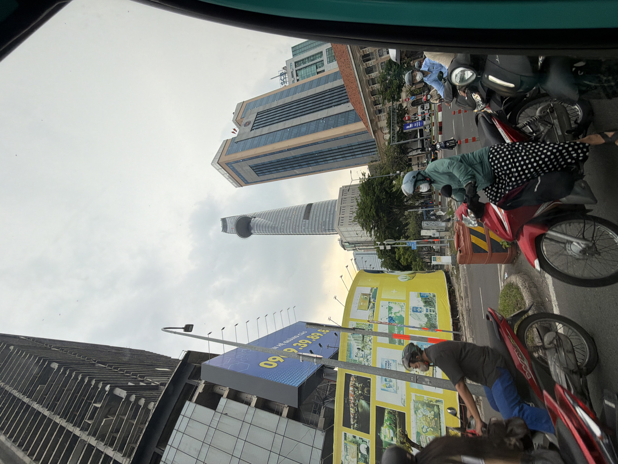 The image captures a street scene in Thủ Đức, Vietnam, characterized by its urban environment and significant architectural structures. In the foreground, several individuals are riding motorcycles. One person, wearing a blue shirt and helmet, is on a dark motorcycle. Next to them, a person in a green jacket and a white helmet is on a red motorcycle, adorned with a black and white polka dot skirt. Another individual in a dark shirt and helmet is riding a red motorcycle on the left.
The background is dominated by a cluster of modern high-rise buildings. The most prominent is a tall, slender skyscraper with a distinctive circular feature at its apex, reminiscent of Bitexco Financial Tower. Adjacent to it is another imposing building with a beige facade and a grid of blue windows. Further to the right, a glass-fronted building with multiple levels is visible. In the middle ground, a large yellow billboard with various images and text is displayed, alongside a construction site with skeletal frameworks of buildings. Traffic lights and street lamps are also present, indicating a busy urban thoroughfare. The sky is overcast, contributing to a diffused lighting condition. The image is taken from a slightly elevated perspective, possibly from a vehicle.
