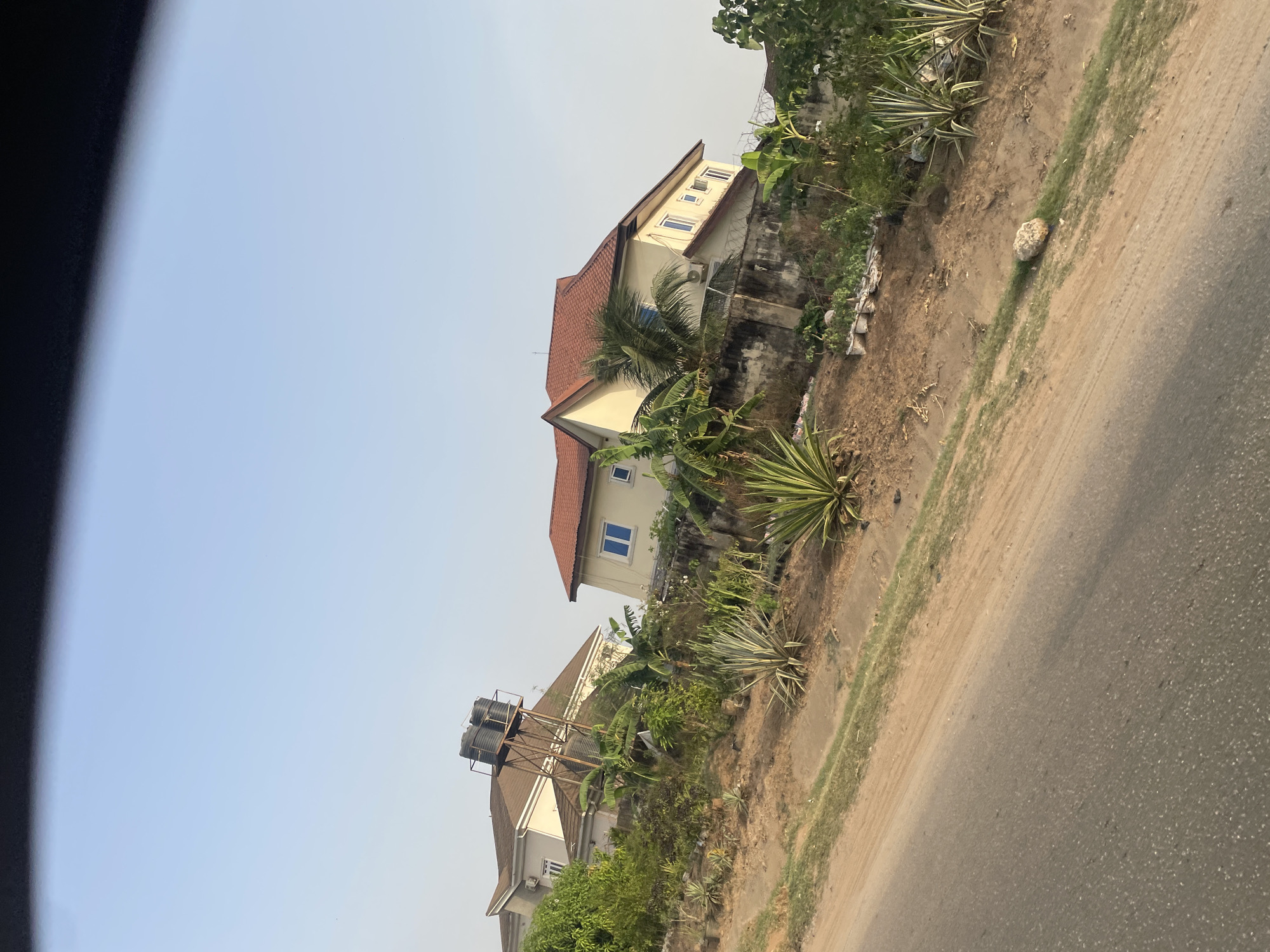 The image depicts a street scene in Galadimawa, Nigeria. Two residential buildings are visible on an elevated plot of land, with red tile roofs and light-colored walls. The first building features a water tower with two black tanks. The second building has a section with multiple small windows. Vegetation, including banana trees and spiky plants, grows along the edge of the property. A dirt shoulder separates the property from an asphalt road. A rock sits on the dirt shoulder. The sky is clear and light blue. The image appears to have been taken from a moving vehicle.