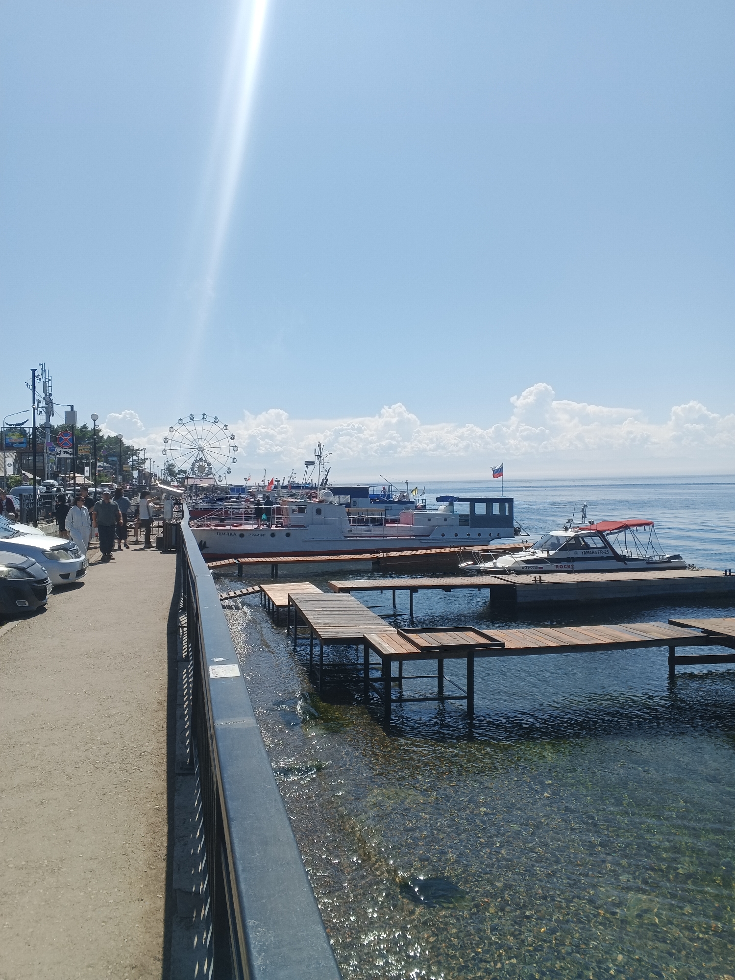 The image depicts a sunny day in Listvyanka, Russia, a popular tourist destination on the shore of Lake Baikal. In the foreground, a paved walkway runs along a metal railing, separating it from the clear, shallow water. Wooden piers extend into the water, with a small white and red motorboat docked at one of them.
Further back, a large white passenger boat is moored at a pier, its hull bearing the Cyrillic inscription "ШИЛКА" (Shilka). Behind the boat, a Ferris wheel is visible in the distance, a common attraction in tourist towns. Several other boats and structures are also present along the waterfront.
On the walkway, a group of people are walking and standing, some appearing to be tourists. Cars are parked along the street on the left side of the image. The sky is a clear blue with a few fluffy white clouds, and a bright sunbeam cuts across the upper portion of the image. The overall scene conveys a sense of a vibrant, sunny day at a lakeside resort.