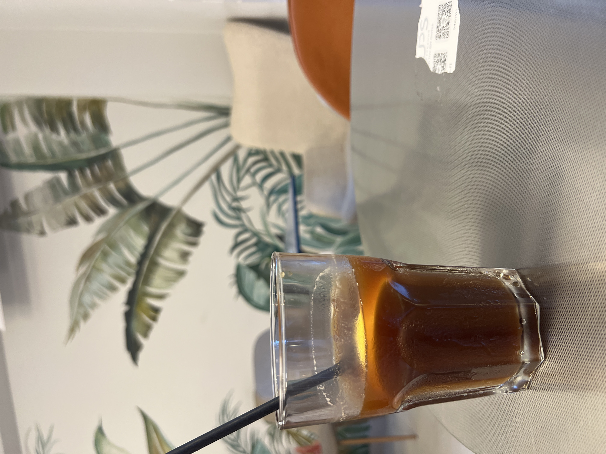 A close-up shot captures a glass of iced coffee with a straw on a textured, reflective tabletop in Budva, Montenegro. The beverage is a deep amber color, and a slice of lemon or orange floats on top. The glass is sweating, indicating it's cold. In the blurred background, a wall is decorated with a tropical mural featuring large green leaves. Two chairs, one a light beige armchair and another with an orange seat, are visible behind the table, along with hints of other patrons. A small receipt or label with QR codes is partially visible on the tabletop to the right. The overall ambiance suggests a relaxed setting, likely a cafe or restaurant.