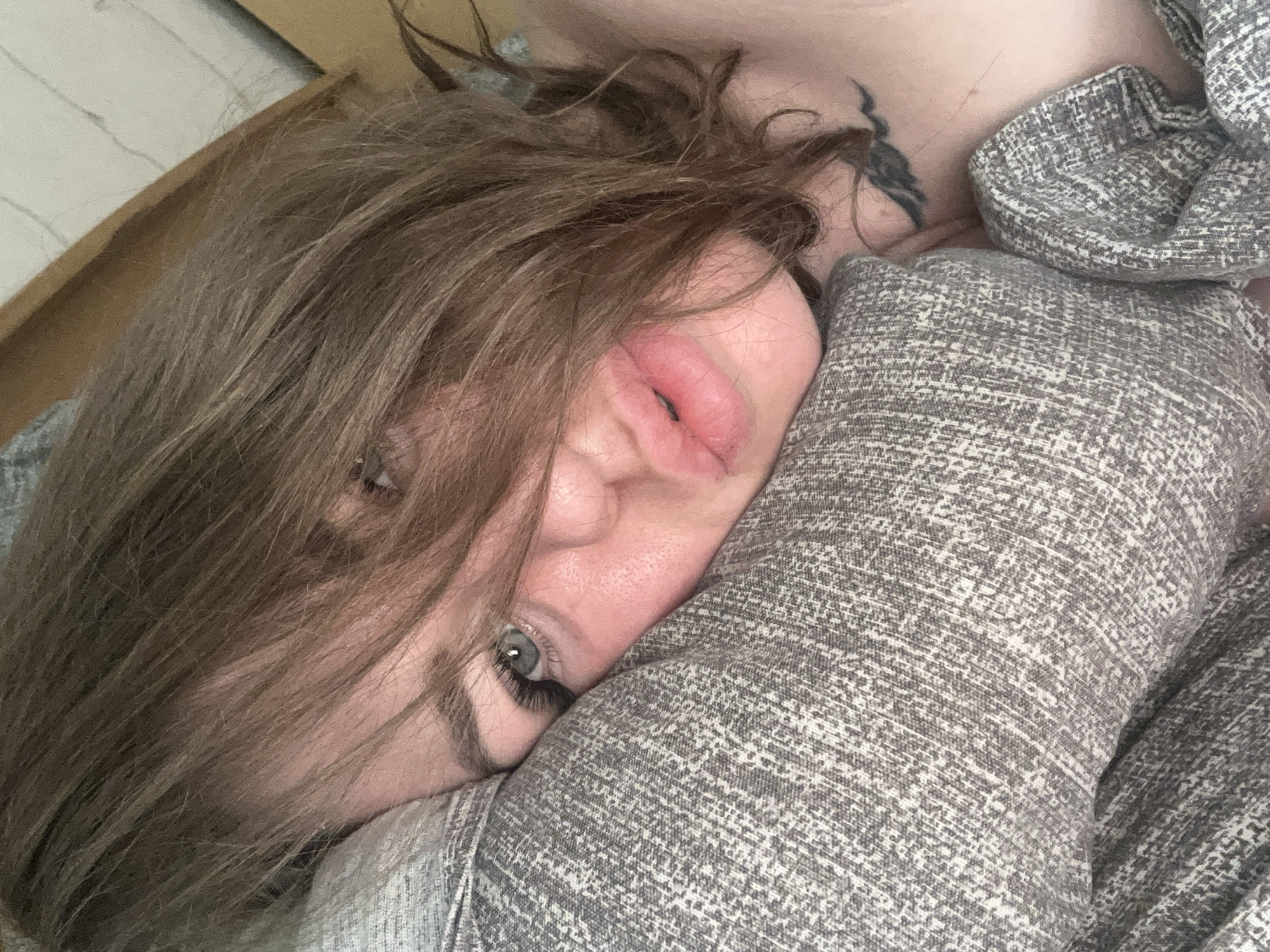 A medium shot captures a young woman from the chest up, lying down on a textured gray pillow. Her head is tilted to her left, and her face is partially obscured by strands of light brown hair. Her left eye, with long lashes and visible eyeliner, is open and looking towards the camera. Her lips are slightly parted, revealing a hint of her teeth. A small, black wing tattoo is visible on her right collarbone. The background is out of focus, suggesting an indoor setting. The lighting is soft and natural.