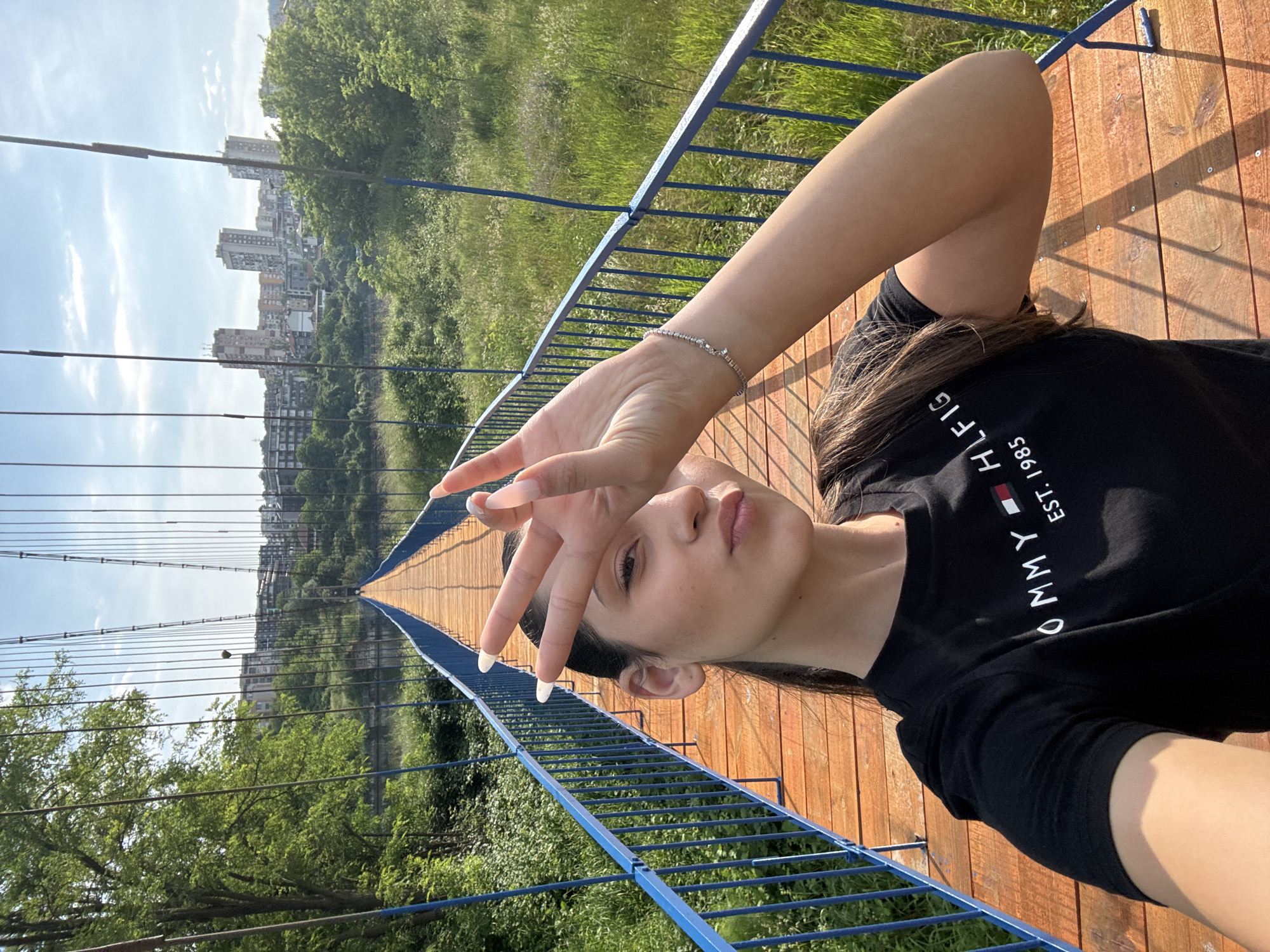A young woman is captured in a selfie taken on a sunny day, likely on a bridge. She is positioned in the foreground, looking directly at the camera with her lips slightly parted. Her right hand is raised, making a "peace sign" (V-sign) and partially covering her face. She has long dark hair and is wearing a black t-shirt featuring the white text "TOMMY HILFIGER EST. 1985" on the chest. A delicate silver-colored bracelet is visible on her left wrist, and her fingernails have distinct white tips.
The woman is lying or leaning on the light brown wooden planks of a bridge deck. The bridge has blue metal railings and visible suspension cables that stretch into the distance. Below the bridge and to its sides, a vibrant green landscape of trees and grass is evident. In the far background, under a clear blue sky with some white clouds, a cityscape with several multi-story buildings can be seen, indicating an urban or suburban setting beyond the natural environment. The overall scene suggests a relaxed and pleasant outdoor moment.