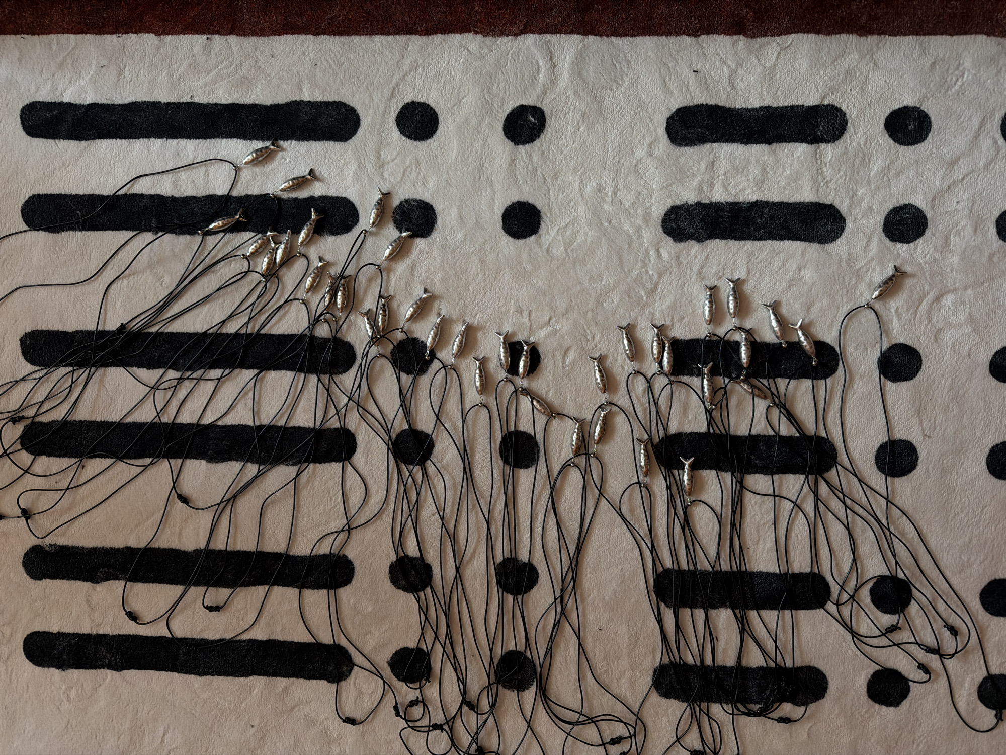 The image shows a cream-colored rug with a pattern of black vertical bars and black dots. Numerous silver-colored fish charms are attached to thin black cords, which are spread out across the rug. The charms are arranged in clusters and lines, with many of the cords tangled. The scene appears to be a close-up, overhead view of the rug and the fish charms. The image does not provide any information about the city of Khimki, Russia, or any people, actions, or interactions.