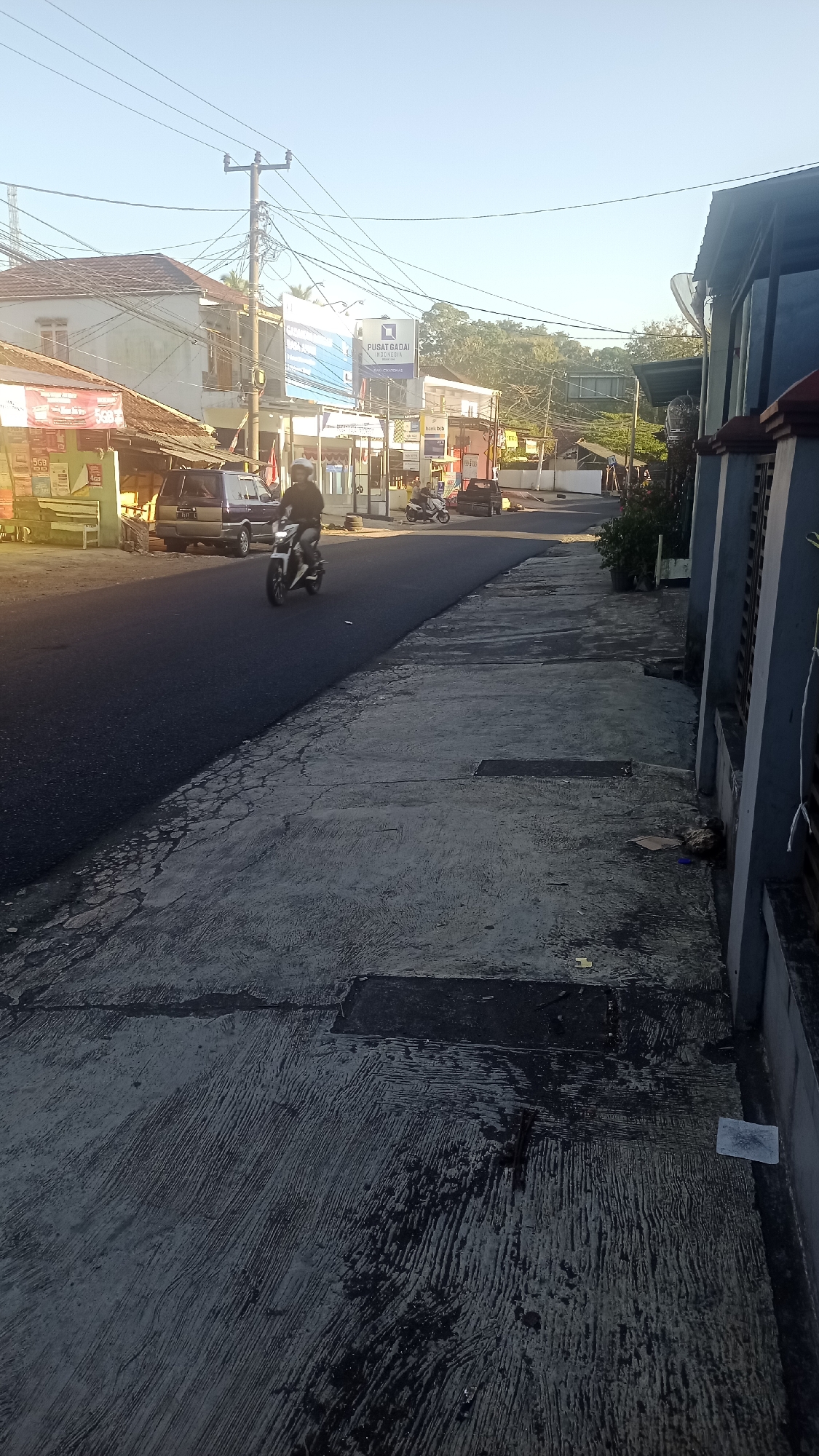 The image depicts a street scene in Pakemitan, Indonesia, bathed in the warm light of what appears to be early morning or late afternoon. The perspective is from a sidewalk looking towards a street.
On the left side of the street, a row of buildings is visible. One building has a weathered, light-colored facade with a red-tiled roof. Adjacent to it, a lighter-colored structure features a prominent blue and white billboard with text in Indonesian, including "PUSAT GADAI INDONESIA". Several smaller storefronts or stalls are also present, one with a red awning and another displaying advertisements. A dark-colored car is parked on the side of the road, and a motorcycle with a rider is in motion on the street. The rider, wearing a dark jacket and a white helmet, is facing away from the camera and appears to be accelerating. Further down the street, another motorcycle is visible.
The street itself is paved and appears to be asphalt. A sidewalk runs along the right side of the image, composed of concrete slabs with a textured, grooved finish. The sidewalk shows signs of wear and tear, with some cracks and darker patches. Debris, including pieces of paper and what appears to be a plastic bag, are scattered on the sidewalk.
In the background, trees and foliage are visible, suggesting a semi-urban or suburban environment. Power lines crisscross the sky, supported by utility poles. The sky is clear and blue. The overall impression is of a typical street in a residential or commercial area of an Indonesian town.