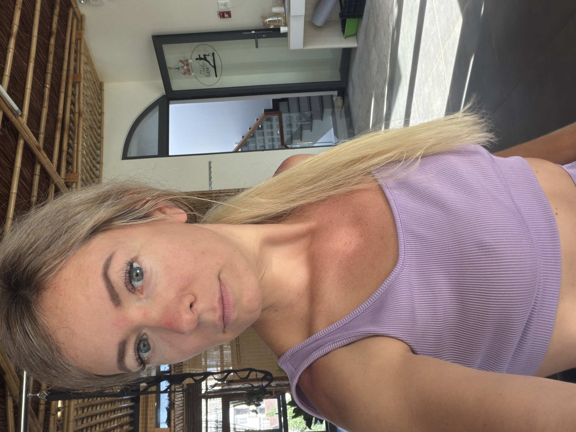 A young Caucasian woman with blonde hair tied in a ponytail is taking a selfie in a yoga studio. She is wearing a purple ribbed tank top and appears to be looking directly at the camera with a neutral expression. The studio has a natural, rustic aesthetic with a bamboo ceiling. In the background, there is a glass door with a logo that reads "Dalat Gem," indicating the location. A staircase is visible through the door, and there are yoga mats and equipment in the studio. The floor is tiled and reflects sunlight, creating dramatic shadows. The scene suggests a bright, airy, and well-lit space, possibly a fitness or wellness center.