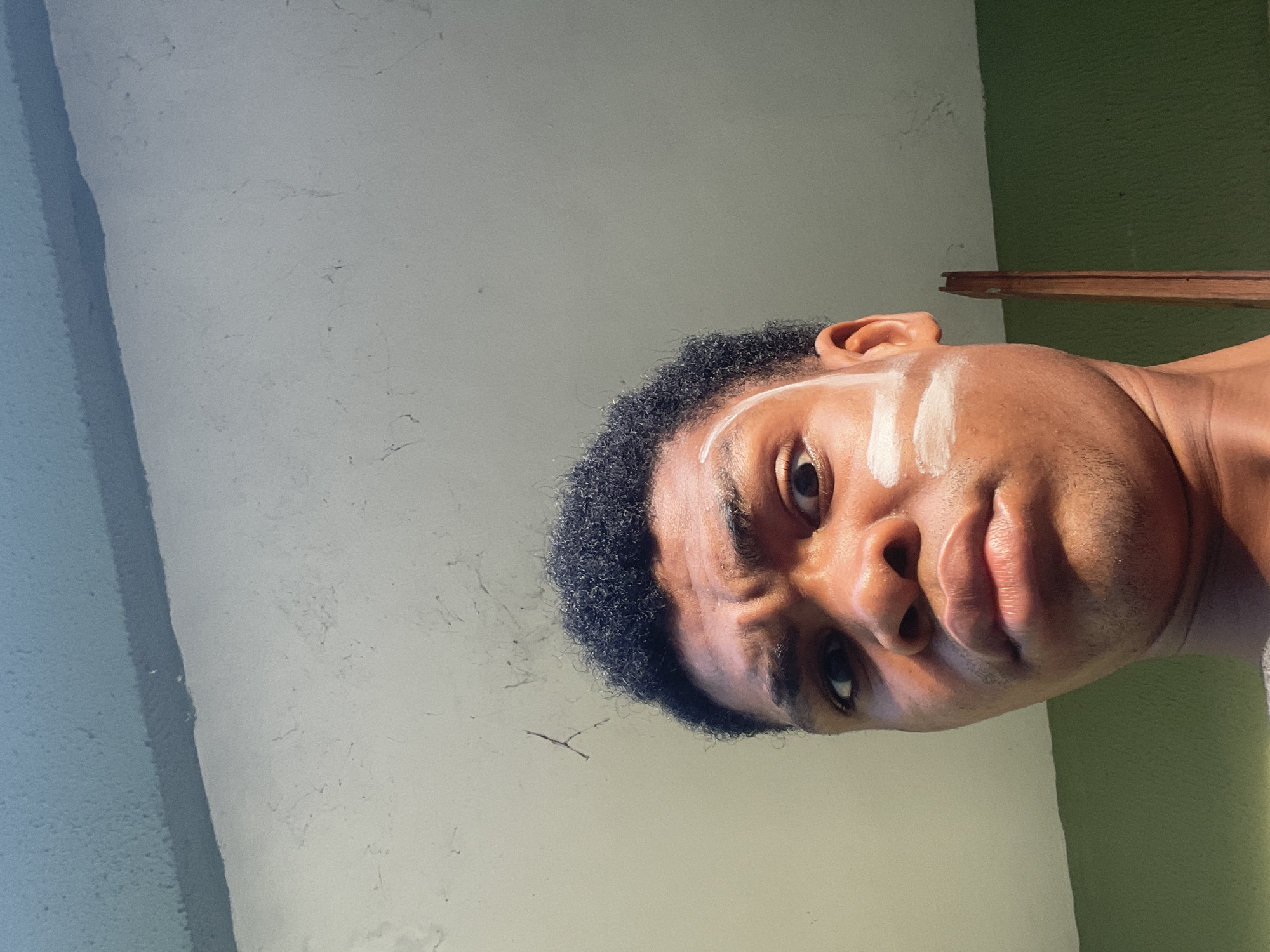 The image is a close-up, rotated portrait of a dark-skinned male. He has short, dark, curly hair. Two vertical white streaks are painted on his forehead above his left eye. The background is a light-colored wall with some texture, transitioning to a green wall on the right. A thin, dark wooden beam is visible partially on the right edge of the image. No information is available about the location of the image in Ado, Nigeria.