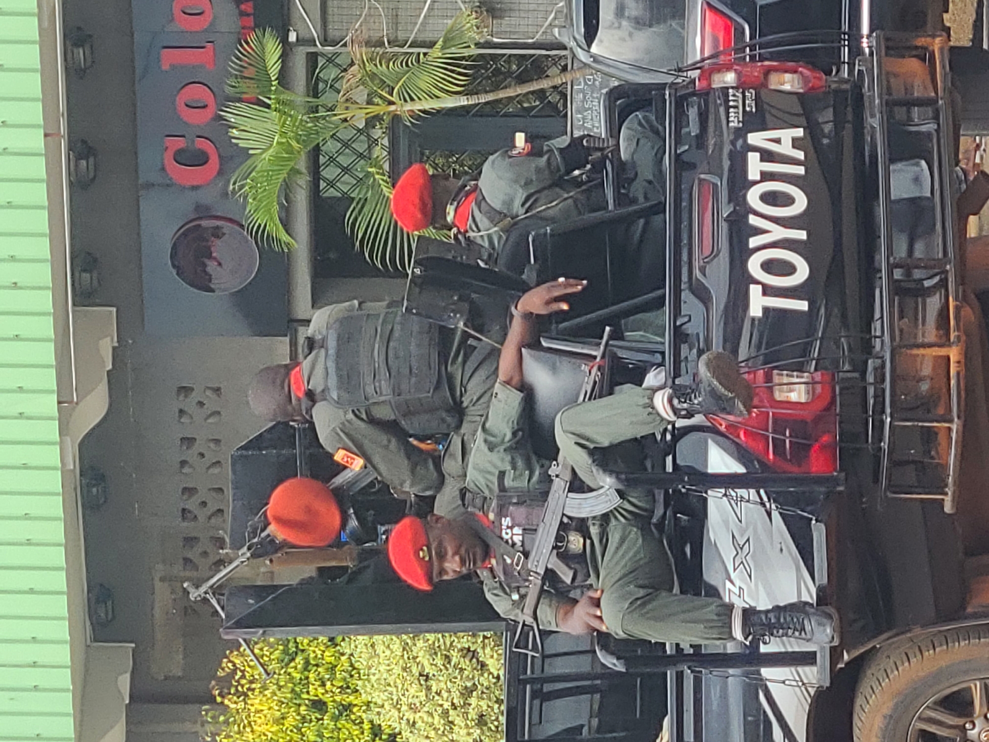 Three uniformed men are seated in the bed of a black Toyota Hilux pickup truck, which is stopped on an urban street. The man in the foreground, seated on the left, is of medium complexion and wears a red beret, an olive green short-sleeved uniform, and a dark tactical vest with "IG'S COMP" visible on it. He holds an AK-pattern rifle across his lap and rests his hands on it and his left leg. He wears black tactical boots with white socks. Behind him, a second man, also wearing a red beret and an olive green uniform with a dark tactical vest, is seated facing away from the viewer, with a "33" patch on his right arm. To the right, a third man with a red beret and olive green uniform is seated facing away.
A heavy machine gun, possibly a DShK, is mounted on a stand behind the second man, with an ammunition belt visible. Additional rifles are positioned upright near the mounted machine gun. The pickup truck's tailgate displays "TOYOTA" in white capital letters and "HILUX" and "SR5" in smaller text. The truck's side shows "FX4" branding.
In the background, a building with a light green corrugated metal roof is visible. Below the roof, a dark gray facade features a black sign with red letters partially spelling "Colo." A window with a diamond-patterned grille is below the sign. Green foliage and a small palm tree are present on both sides of the background. The scene is located in the city of Unknown, Unknown.