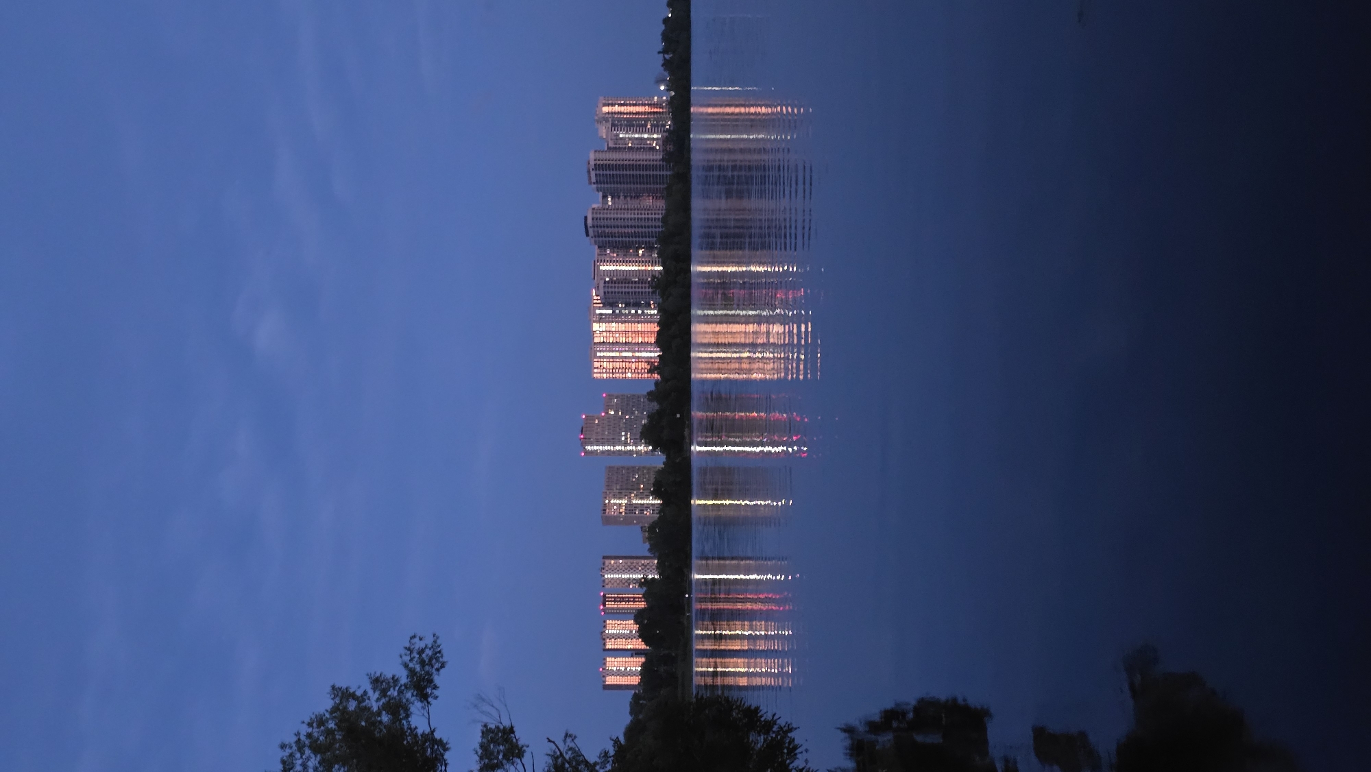 This is a vertical, eye-level, full shot photograph taken at dusk of apartment buildings reflected in a body of water in Kyiv, Ukraine. The sky is a deep blue with wispy clouds, and the water is a dark, reflective surface. A tree branch with dark green leaves is visible in the upper left corner, partially obscuring the sky.
In the background, across the water, a line of tall, modern apartment buildings stretches horizontally across the frame. The buildings are illuminated from within, with windows glowing in various shades of orange and white, suggesting that residents are home. The lights from the buildings are prominently reflected in the water below, creating a mirror image of the cityscape. The buildings appear to be part of a modern residential development.
The water itself is calm, with gentle ripples disturbing the perfect reflection of the buildings. The overall mood is serene and tranquil, despite the urban setting. The time of day, indicated by the dusk sky and illuminated windows, suggests an evening scene. There are no people or vehicles visible in the frame, focusing the composition entirely on the natural environment and the architectural landscape. The location is implied to be a waterfront area within Kyiv, Ukraine, possibly a river or a large lake that borders a residential district.